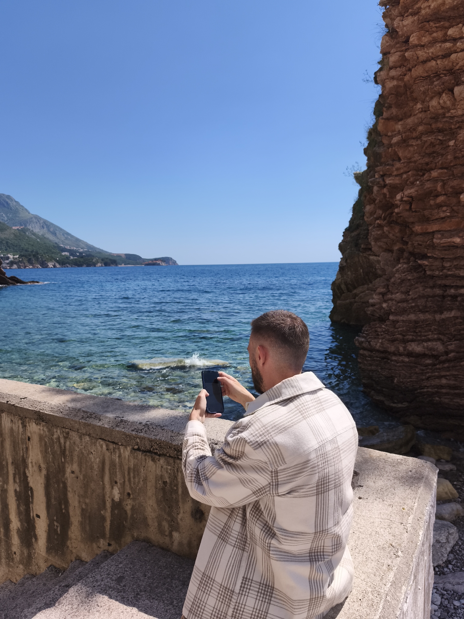 The image depicts a man in a plaid shirt taking a photo with his smartphone of the Adriatic Sea and the coastline near Rafailovići, Montenegro. He is standing on a concrete ledge or low wall near a rocky outcropping. The water is clear and turquoise, and the sky is a bright blue. Mountains and coastal settlements can be seen in the distance.