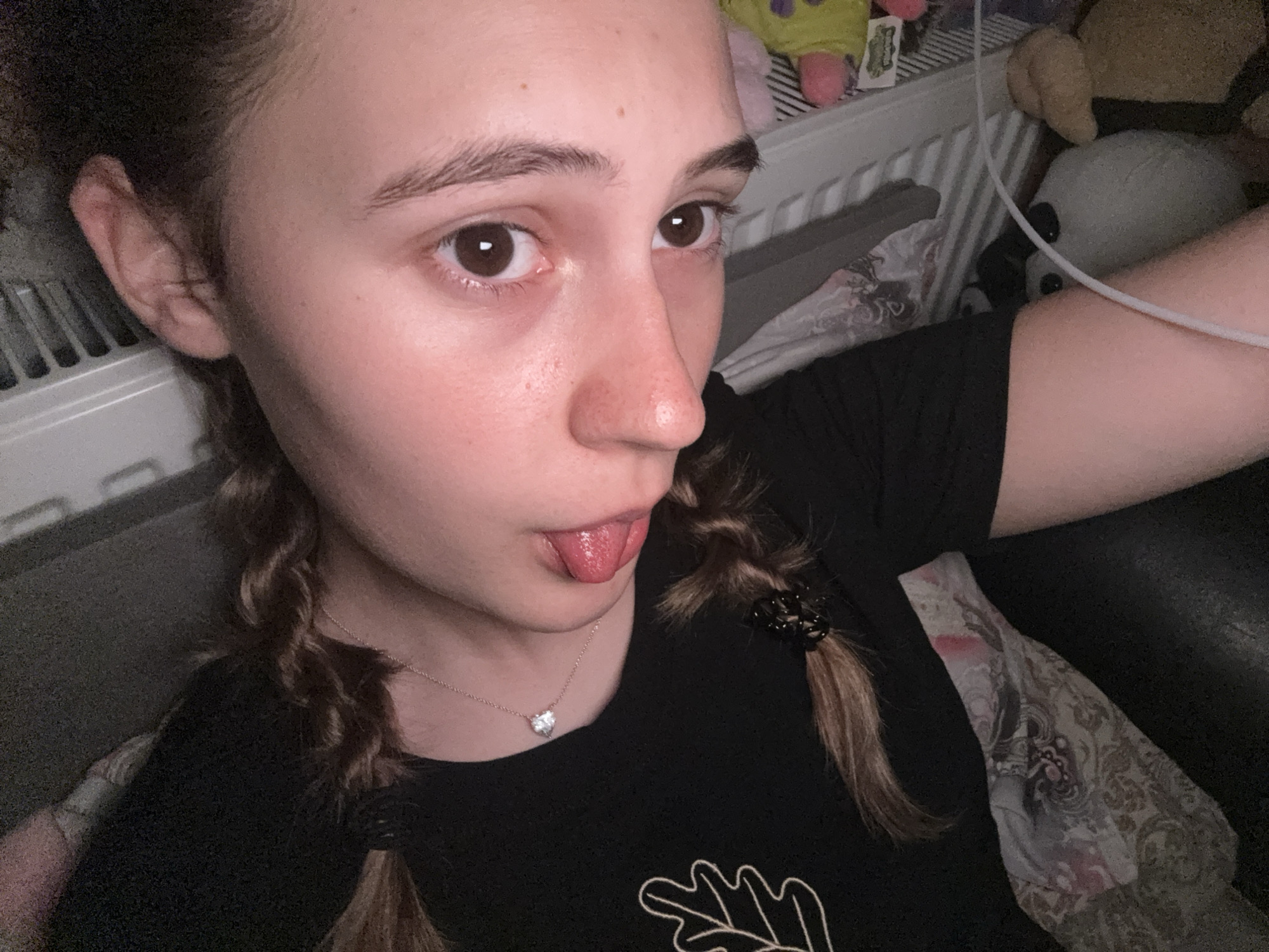 This close-up selfie captures a young individual, likely female, in an indoor setting in Panagyurishte, Bulgaria. The subject has fair skin, prominent dark brown eyes, and dark brown hair styled into two braids, each secured with a black elastic tie. She is wearing a black short-sleeved t-shirt with a light-colored, partial outline of a design, possibly a leaf or antler, visible on the chest. A silver necklace with a small, heart-shaped pendant adorns her neck.
The person is looking slightly to the right of the camera and playfully sticking out her tongue, conveying a lighthearted and casual mood. The background indicates a personal space, likely a bedroom or living area. A white radiator is visible behind her left ear. To the upper right and behind her right arm, several plush toys are partially visible, including what appears to be a white panda. A thin white cable runs diagonally across the upper right portion of the frame. Parts of a patterned fabric, possibly a blanket or cushion, are also seen. The lighting is soft and ambient, characteristic of an indoor environment, potentially in the evening. No specific weather conditions are discernible from the image.