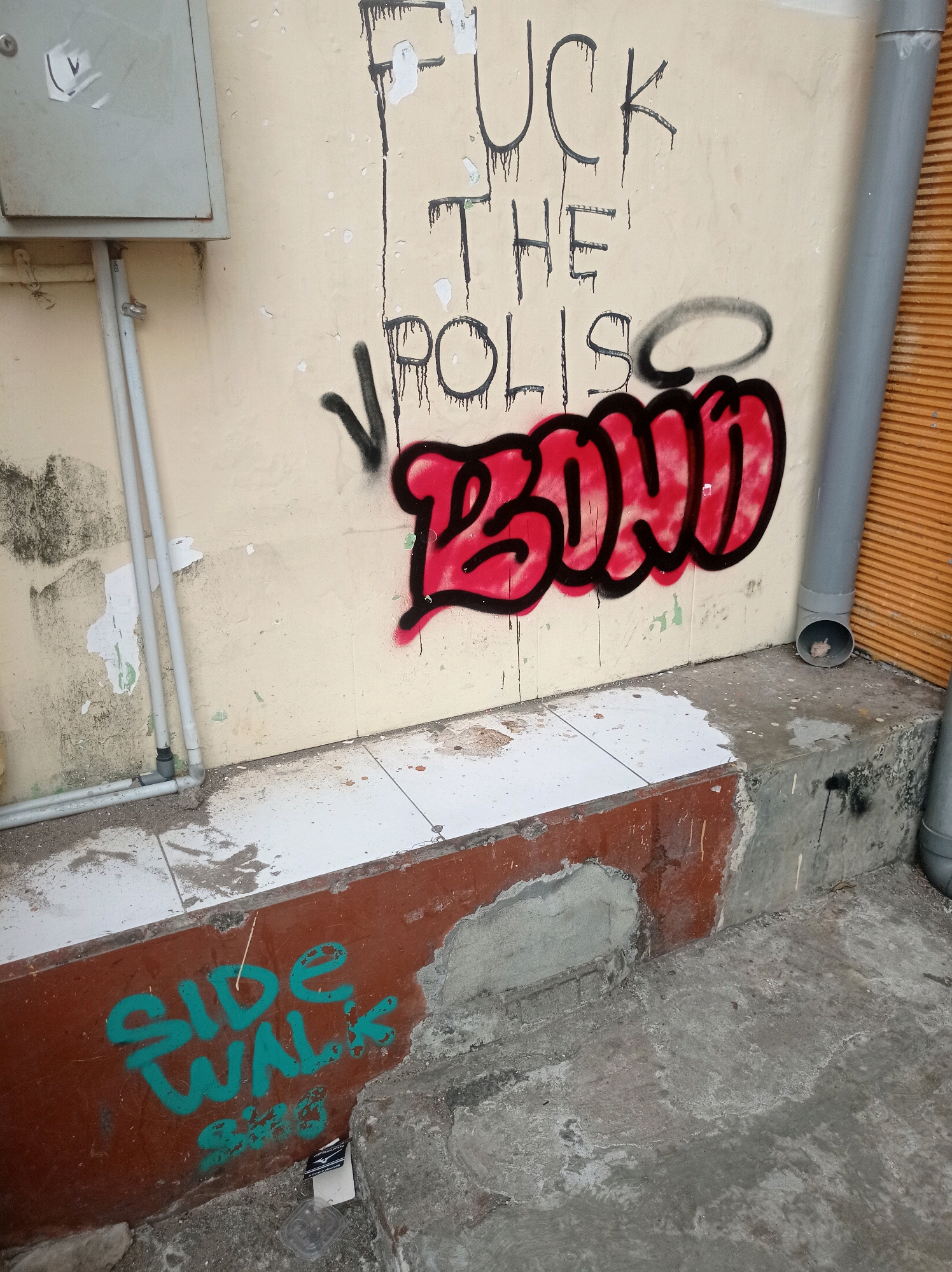 In Tuban, Indonesia, graffiti mars a light-colored building. The graffiti, rendered in black spray paint, reads "FUCK THE POLIS" vertically down the wall, with "POLIS" partially obscured by a red spray-painted tag that appears to say "BOOO". Below this, in teal spray paint on a reddish-brown section of the wall, are the words "SIDE WALK" with smaller, illegible text beneath it. The scene also includes electrical conduits, a drainpipe, and a concrete walkway.