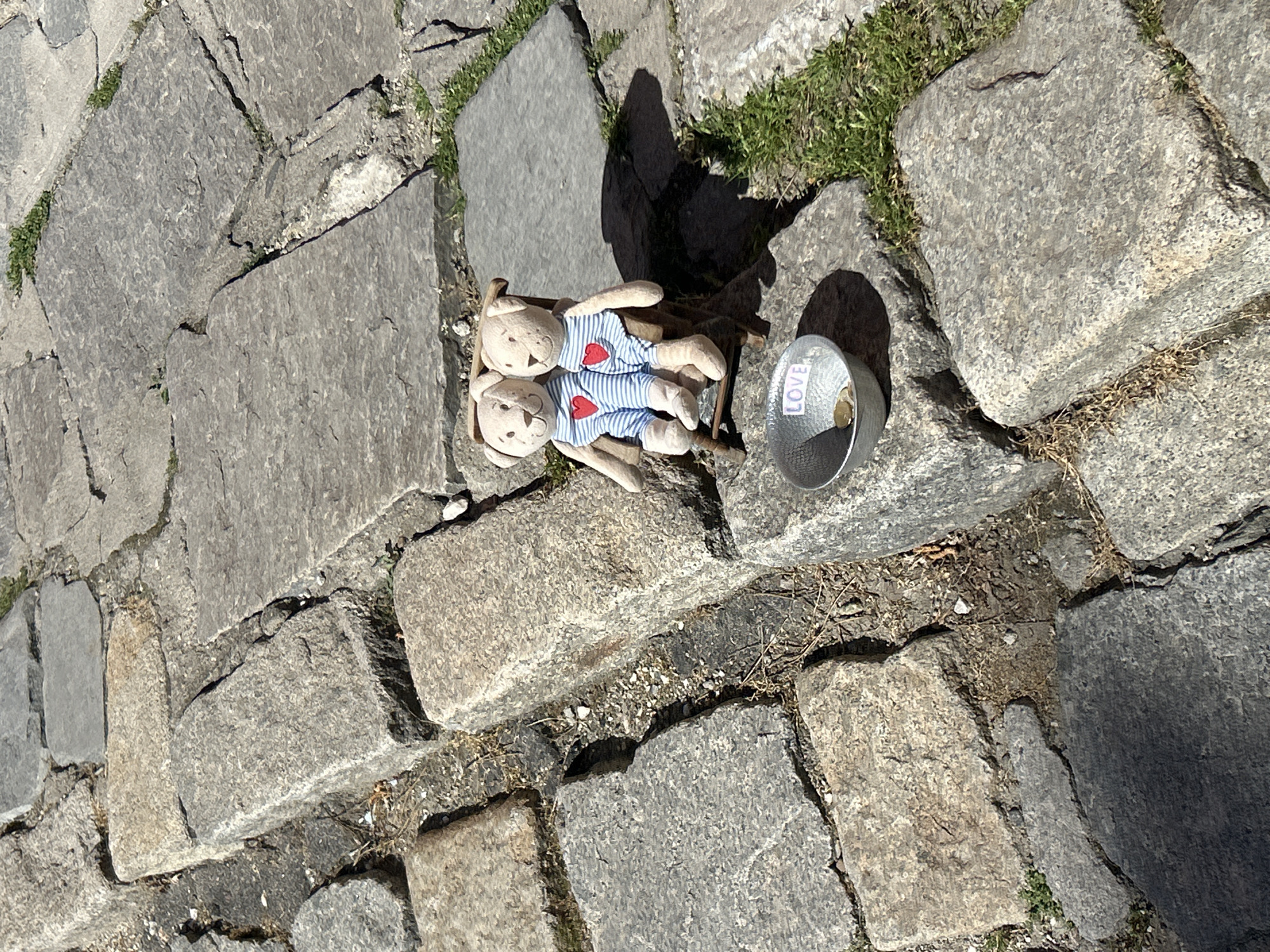Two stuffed bears wearing blue and white striped shirts with red hearts lie on a small wooden sled on cobblestones. Next to them is a small, silver-colored metal cup containing coins and a piece of paper that says "LOVE". The cobblestones are gray and unevenly shaped. Grass grows between some of the stones. The scene is likely outdoors, judging by the lighting and the cobblestone ground. Based on the style of cobblestones and the potential for such an arrangement, this could be a street scene in Plovdiv, Bulgaria.