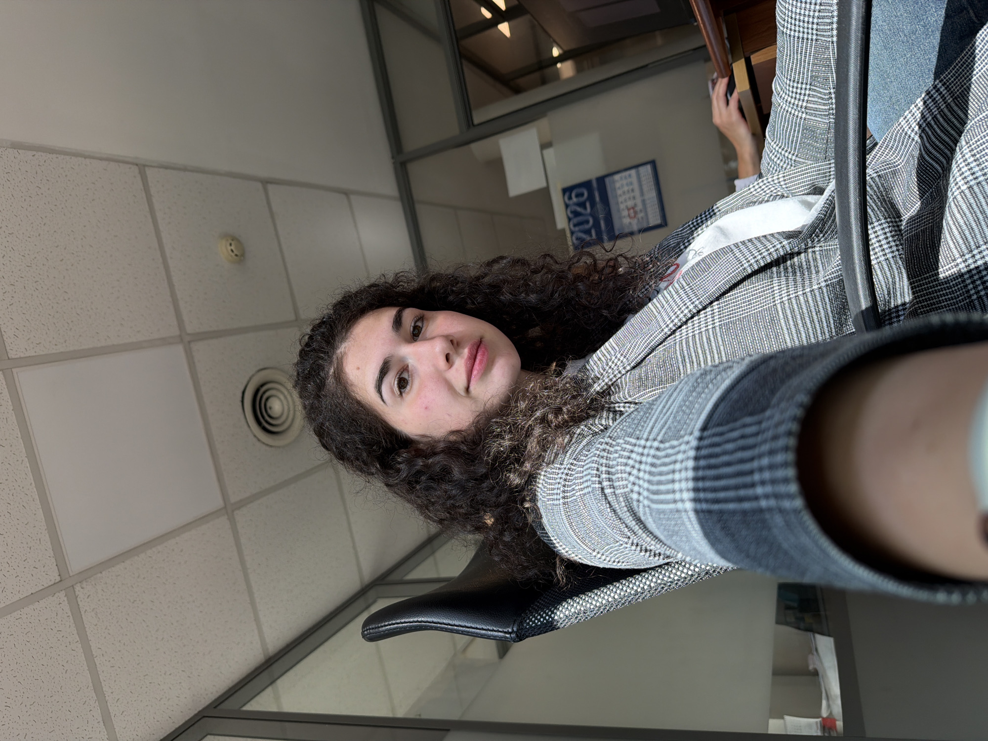 In this first-person perspective image, a young woman with dark, curly hair is seated at a desk in an office environment. She is wearing a grey and white plaid blazer over a white t-shirt and blue jeans. Her right arm is extended towards the camera, obscuring part of the foreground. Her left hand is on a computer mouse, with her fingers positioned to click. In the background, a calendar with the year "2026" is visible on the wall. The room features a tiled ceiling with fluorescent lighting and glass partitions. The overall setting suggests a modern office space within the city of Sofia, Bulgaria.