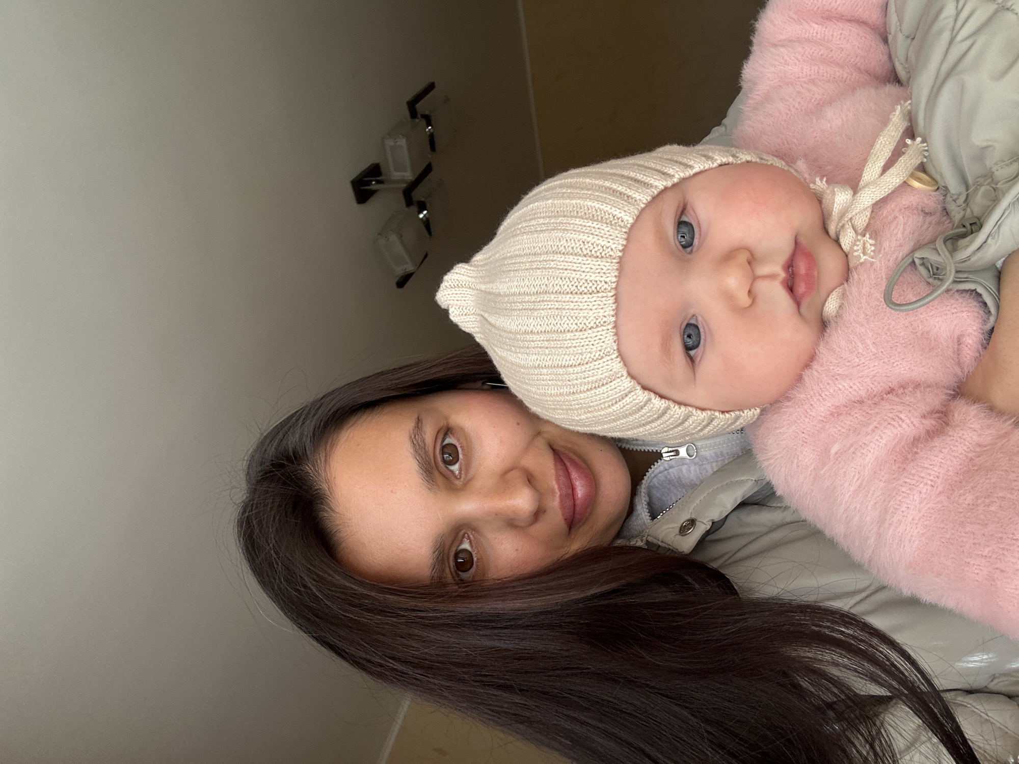 A woman and baby are in an indoor setting. The woman has dark brown hair, brown eyes, and fair skin. The baby has fair skin, blue eyes, and is wearing a knitted cap. The baby is wearing a pink garment. The woman is wearing a grey jacket. The background shows a wall with a light fixture. There is nothing to specifically identify Chaikovsky, Russia as the location.
