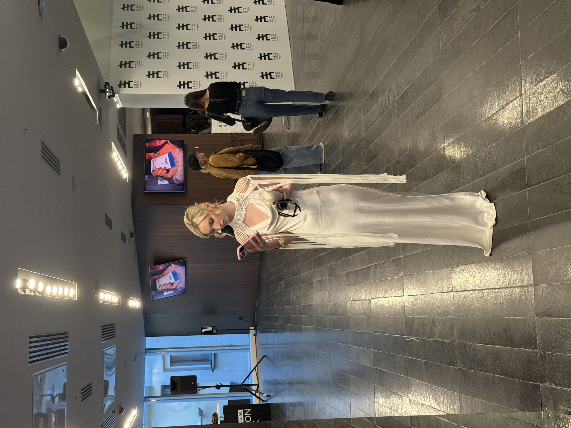 The image, rotated 90 degrees clockwise, captures an indoor event or gathering in what appears to be a modern venue. In the foreground, a woman with blonde hair is prominent, wearing an elegant, floor-length white dress with intricate, possibly beaded or woven, detailing around the neckline and shoulders. She is engrossed in her pink smartphone, looking down at the screen.
In the midground, two other individuals are visible from behind. One person wears a brown jacket over a dark top and light-wash jeans, carrying a dark bag. The other is dressed in a black top and blue jeans.
The background features a white wall adorned with a repeating black logo or pattern. This pattern includes the Cyrillic text "НОВАЯ СЦЕНА," which translates to "New Stage," and a stylized "HE." Several flat-screen monitors are mounted on the walls; one screen displays content with the partially visible text "BAPTREP." The floor is covered with dark, rectangular tiles laid in a diagonal pattern, reflecting some of the artificial light.
The space is well-lit by multiple rows of spotlights recessed into the ceiling. In the lower left, a black speaker on a stand is visible, with text that appears to read "SBURG ON," likely referring to Saint Petersburg, Russia, aligning with the provided location context. The scene suggests a sophisticated environment, possibly a pre-show reception, gallery opening, or a similar cultural event at the "New Stage" in Saint Petersburg. The individuals seem to be mingling or waiting.