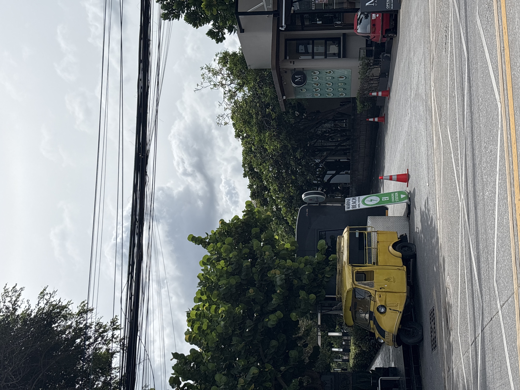 The image captures a street scene in Pattaya City, Thailand, on an overcast day. In the foreground, a yellow three-wheeled vehicle, resembling a tuk-tuk, is parked on the asphalt. The vehicle has a covered passenger area and a utility bed at the rear. To the right of the vehicle, a green sign with white lettering reads "MARINE BEACH HOTEL PATTAYA" and "EV CHARGING STATION."
Further back, on the right side of the frame, is a modern building with a gray facade and a glass storefront. A sign above the entrance displays the letter "M" inside a circle, with "CAFE" below it. To the far right, a small red car is parked.
The background features dense green foliage and trees, suggesting a tropical environment. A complex network of overhead electrical wires crisscrosses the upper portion of the image. Several orange traffic cones are placed on the road near the building. The overall impression is a typical urban street in a coastal city, with elements of local transportation and commercial establishments.