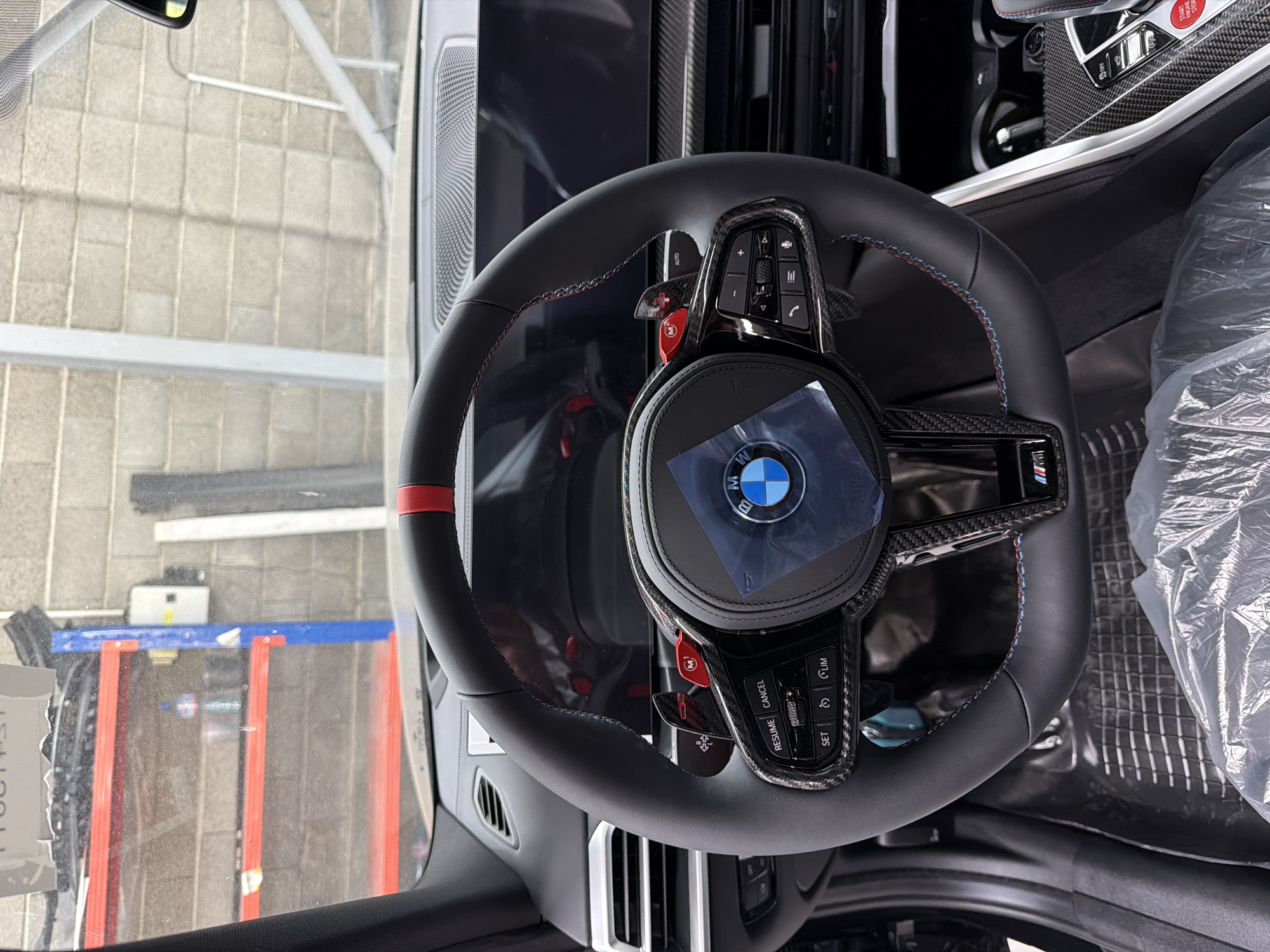 The image shows the interior of a modern car, specifically the steering wheel and dashboard area. The steering wheel is black leather with red stitching and a red stripe at the top. It features various buttons and a central BMW logo. Carbon fiber accents are visible on the steering wheel and surrounding trim. The dashboard includes a large screen, air vents, and other controls. The car appears to be in a storage or workshop environment, with shelves containing items visible in the background through the windshield. The floor is covered with a protective plastic mat. There are no people visible in the image. The location is described as Burgas, Bulgaria, but this cannot be determined from the visual information in the image itself.