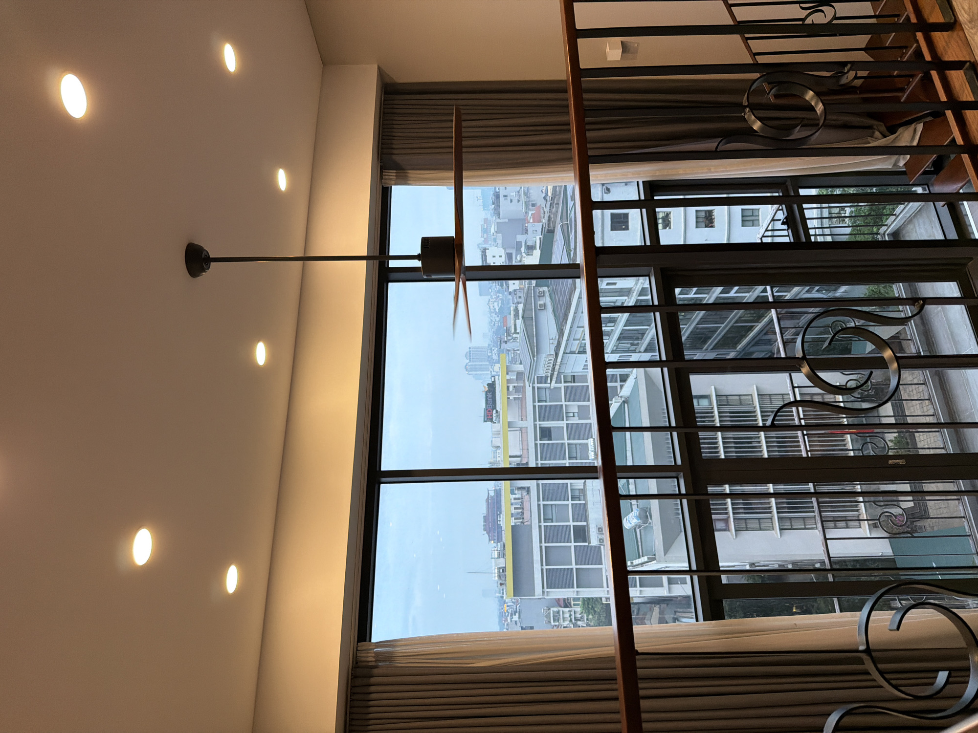 This image shows an interior view looking out a large window at the cityscape of Hanoi, Vietnam. The viewpoint is from a mezzanine or upper level, indicated by the railing in the foreground. The room features recessed ceiling lights and a modern ceiling fan with dark wooden blades, which appears to be in motion. The window is expansive, offering a panoramic view of the city.
The city view consists of various buildings of different heights and architectural styles. There are modern apartment buildings, some with visible balconies and windows, as well as older structures. In the mid-ground, a prominent building with a grey facade and numerous windows is visible. In the distance, taller buildings are discernible, suggesting a dense urban environment. The sky is overcast, contributing to a diffused light.
The interior elements visible include thick curtains on either side of the large window, one light beige and the other darker grey. The railing in the foreground is made of dark wood with black metal accents, featuring decorative scrolled elements. A wooden staircase is partially visible on the right side of the image, leading downwards. The overall impression is a modern residential or commercial interior with a significant urban vista.