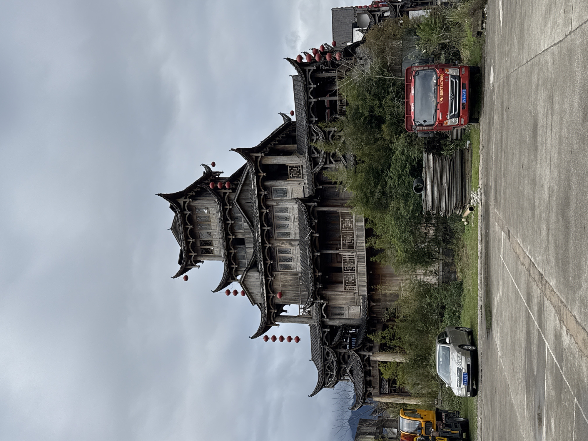 The image displays a multi-tiered wooden building in Wulingyuan, China, under an overcast sky. The structure exhibits traditional Chinese architectural characteristics, featuring multiple dark, curved roofs, intricate wooden balconies, and weathered timber construction. Numerous red spherical lanterns hang from the eaves and balconies across several levels of the building.
In the foreground, a grey concrete lot with faint parking lines and a few shallow puddles occupies the lower half of the frame. A silver Skoda sedan with license plate 湘 A8367 is parked on the left side of the lot, partially on a grassy patch, facing right. To its left, the front portion of a yellow and orange heavy vehicle is partially visible. On the right side of the concrete lot, a red truck with license plate 湘 S7G3K8 and Chinese characters, including a phone number (18807447886), on its front, is parked facing forward, partially obscured by dense green bushes. Behind the red truck, a stack of wooden or metal panels is visible on the ground.
Dense green foliage and some deciduous trees surround the base of the main building and the parked vehicles. Distant, hazy mountains are visible on the far left horizon behind the building. The sky is uniformly grey and cloudy. No individuals are present in the scene.