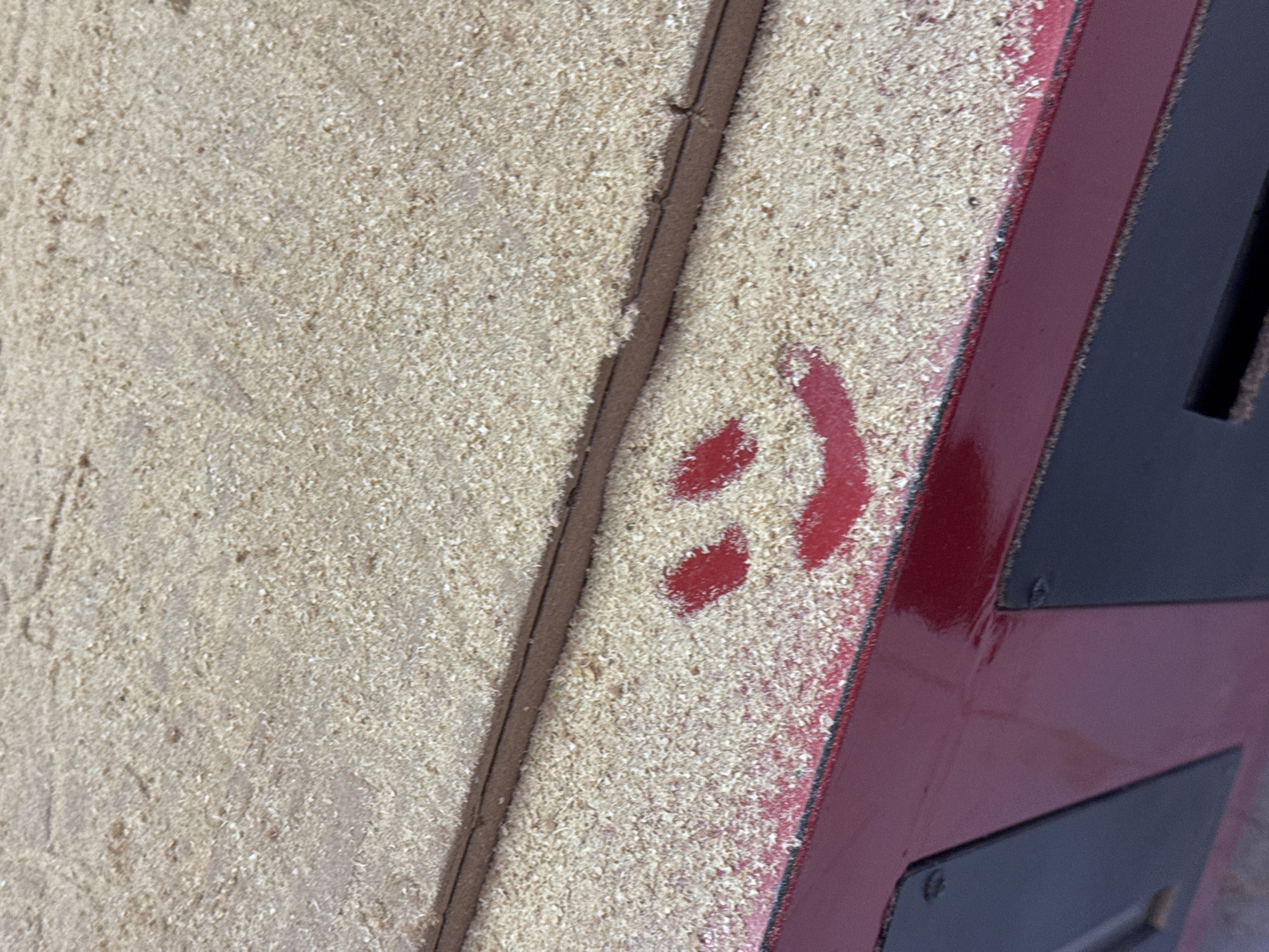 The image shows a close-up view of a surface covered in sawdust. A red, painted surface with what appears to be a stylized ">>" symbol is visible on the right side of the frame. There is a dark trim piece running diagonally from the top left to the lower right. The red surface is glossy and features dark, rectangular elements, possibly panels, attached with screws. The environment is dusty, and the lighting appears to be natural. The city of San Bernardino Tlaxcalancingo, Mexico is not discernible from the image.