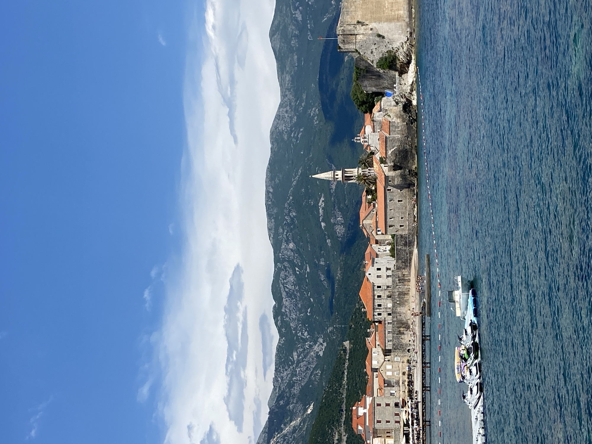 The image depicts the coastal town of Budva, Montenegro, featuring the Old Town, the Adriatic Sea, and the surrounding mountainous terrain. The Old Town is composed of buildings with orange-tiled roofs, punctuated by a prominent church steeple. The town is positioned against a backdrop of rugged mountains, and a segment of a fortified wall is visible near the coastline. The Adriatic Sea occupies the right side of the image, with several boats and docks close to the coast. The sky is blue, with scattered white clouds.