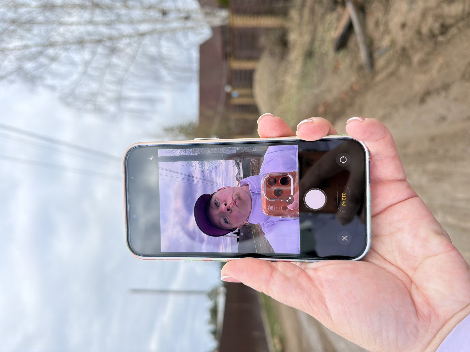 A close-up, vertically oriented outdoor shot (rotated 90 degrees clockwise) features a person's hand holding a modern smartphone, likely an iPhone, enclosed in an orange-pink transparent case. The hand is positioned as if operating the device, with fingers resting on the edges.
The phone's screen displays the live preview of its front-facing camera, capturing a selfie of a young person, possibly a woman or girl. She is wearing a purple baseball cap and a light purple jacket, and is making a playful "fish face" with puckered lips. Her blue eyes are visible, looking directly at the camera. The phone's camera interface is visible around the selfie preview, including a white circular shutter button, a circular icon with arrows for switching cameras, and the text "PHOTO" indicating the current mode. A small "X" icon is also present on the screen.
The background behind the hand and phone shows an outdoor environment in Chaikovsky, Russia. Bare tree branches are visible against a bright, overcast sky, suggesting late winter or early spring. A dirt or gravel path runs into the distance, with indistinct buildings or structures vaguely visible further back. The overall lighting is soft, consistent with an overcast daytime. The visible text on the screen is "PHOTO".