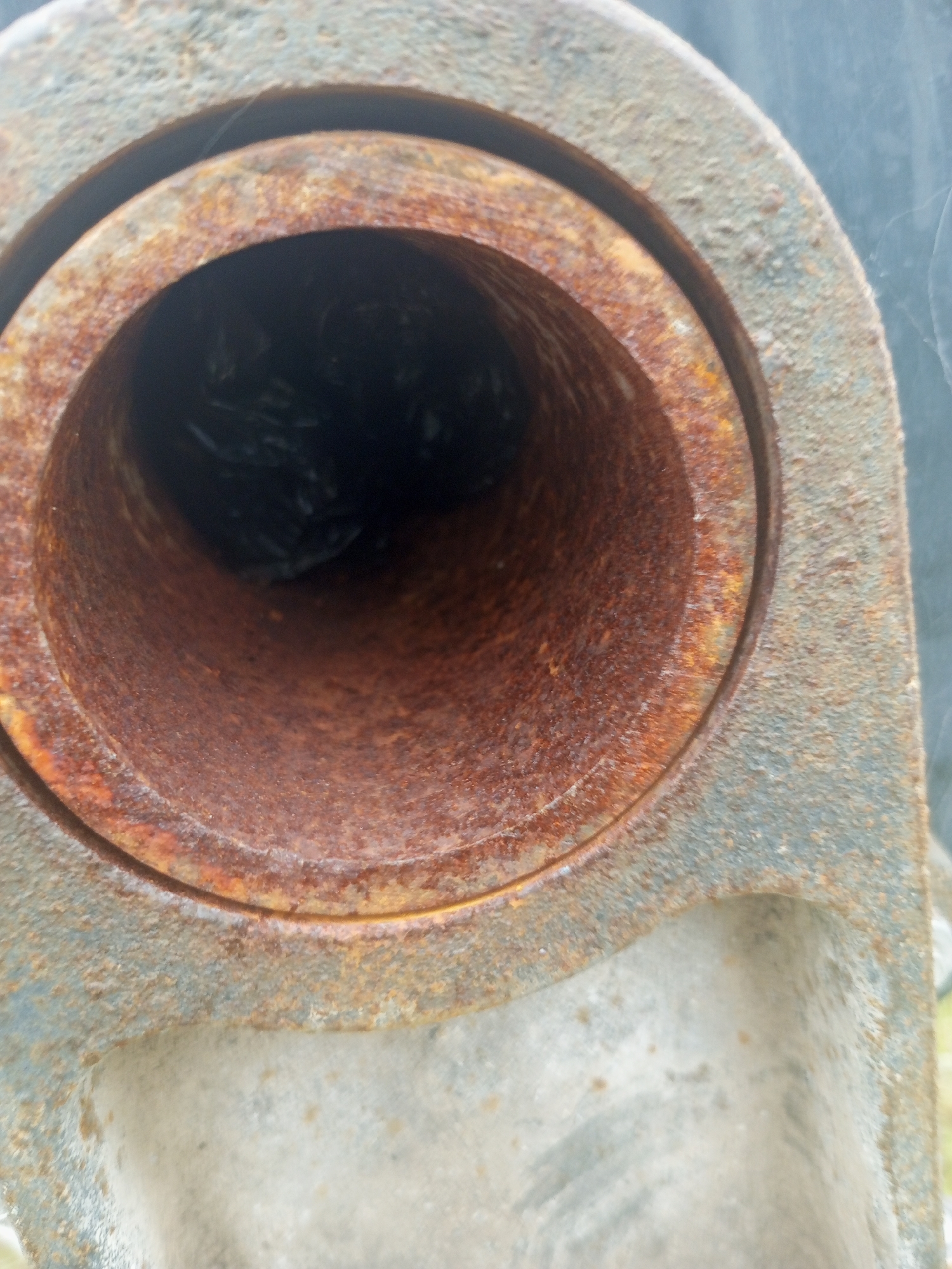 The image shows a close-up view of a rusty, hollow pipe. The pipe is made of metal and has a rough, textured surface with significant corrosion. The interior of the pipe is dark, suggesting it is either empty or filled with something dark and unidentifiable. The exterior of the pipe also shows signs of rust and wear, with some greyish material visible. The focus is on the opening and inner surface of the pipe. There are no people or clear environmental cues in the image, beyond what might be inferred from the texture and material of the object. The lighting appears to be natural, but there are no strong shadows or indications of a specific time of day or weather conditions. The overall impression is one of age and disuse. There is no visible text in the image.