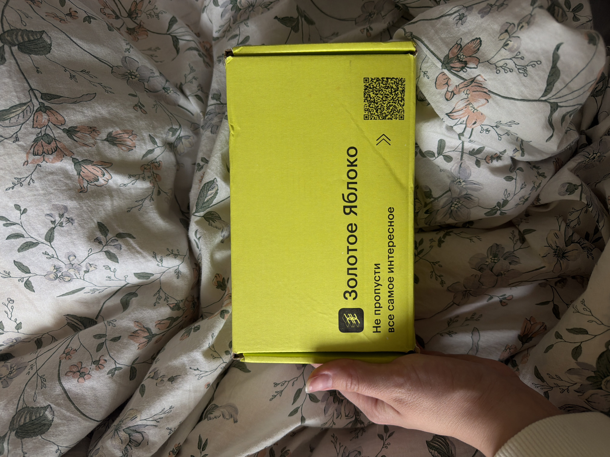 The image shows a yellow box with Russian text that reads "Золотое Яблоко," which translates to "Golden Apple." Below this, the text says "Не пропусти все самое интересное," translating to "Don't miss the most interesting." The box also has a QR code and arrow symbols. A person's hand is partially visible near the bottom right corner of the box. The box is placed on a bedspread with a floral pattern. Based on the brand "Golden Apple," which is a Russian beauty retailer, and the use of Russian text, the image is likely from Russia. However, there is no definitive information in the image to confirm the specific location of Izhevsk.