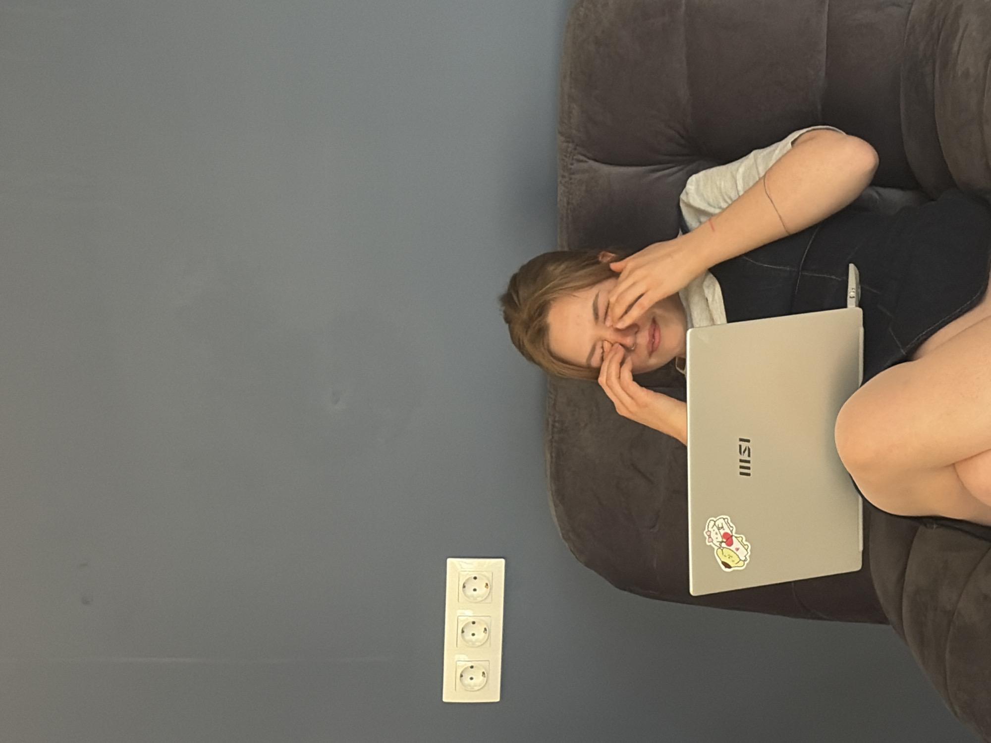 The image shows a person sitting on a gray couch with a silver MSI laptop on their lap. The person is covering their eyes with their hands, appearing distressed. They are wearing a white t-shirt under a dark denim dress. On the wall, there is a white electrical outlet with three sockets. The background is a solid gray wall. The location is Бараўлянскі сельскі Савет, Belarus.