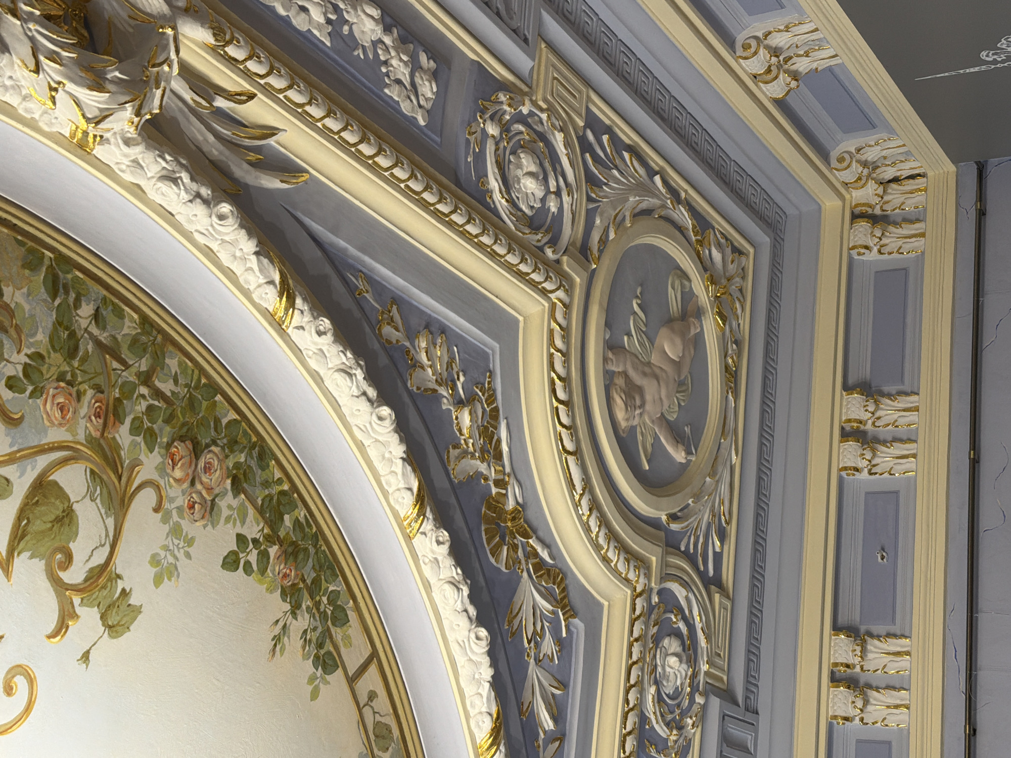 The image shows a detailed view of an ornate ceiling in what appears to be a historical building. The ceiling features intricate plasterwork and gilded details, including floral motifs, scrollwork, and a central oval medallion depicting a cherub. The painting on the upper part of the ceiling depicts roses and greenery. The colors are predominantly pastel shades of blue, cream, and gold, with accents of pink and green in the painted section. The architectural style suggests a neoclassical or baroque influence. This ceiling is likely located in Nizhny Novgorod, Russia, possibly within one of its grand palaces or public buildings. The image is a close-up, vertical shot, focusing on the decorative elements of the ceiling. There are no people or actions visible in the image.
