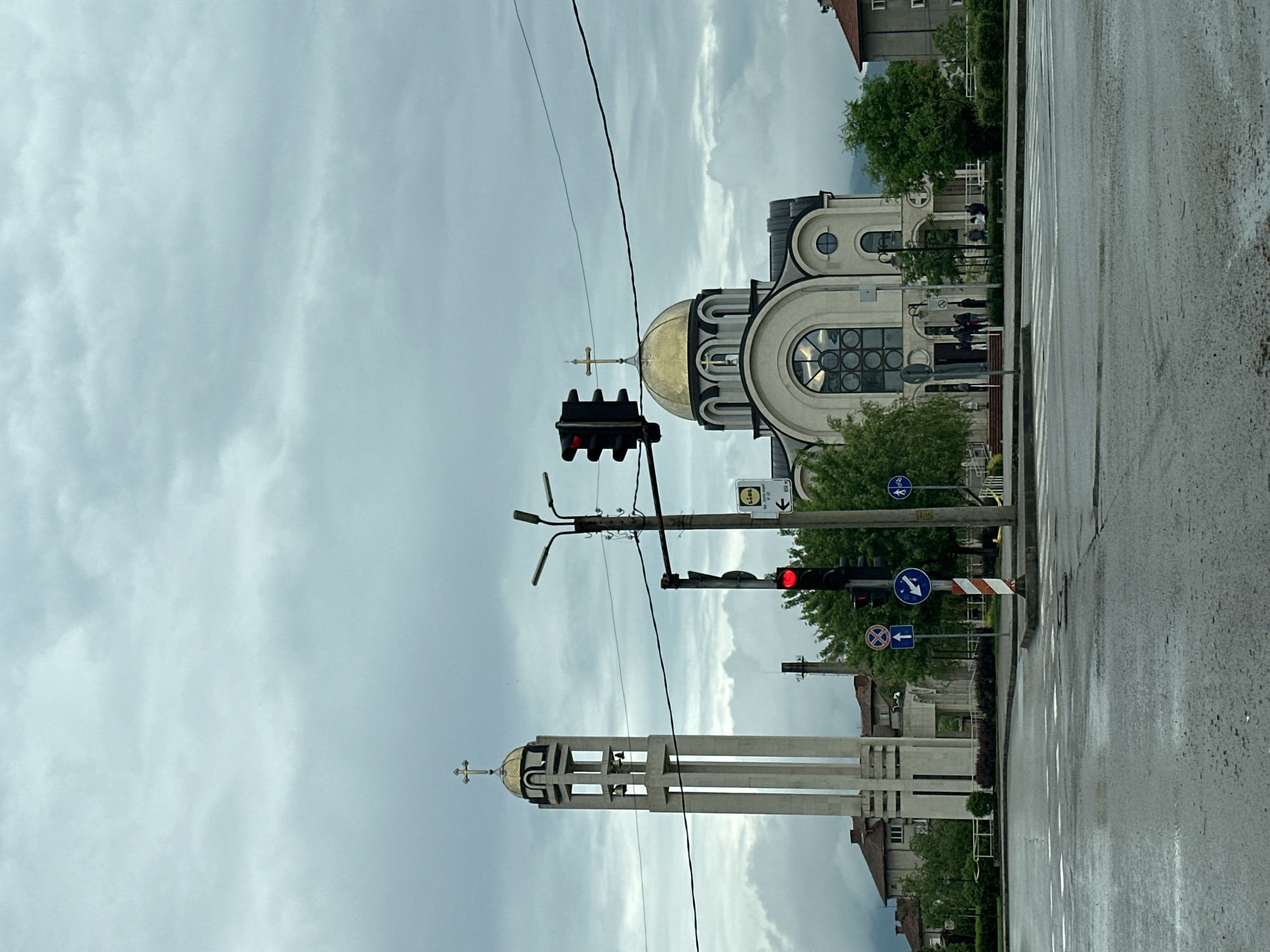 The image shows a street view in Montana, Bulgaria, featuring the St. Cyril and Methodius Orthodox Church. The church consists of two main structures: a tower and a main building with a golden dome, both topped with crosses. A traffic light is visible in the foreground, showing a red light on both its upper and lower heads, with street signs and utility poles nearby. The road appears wet, possibly from recent rain. Buildings and trees line the street, with an overcast sky in the background. People are present on the steps leading to the church entrance.
