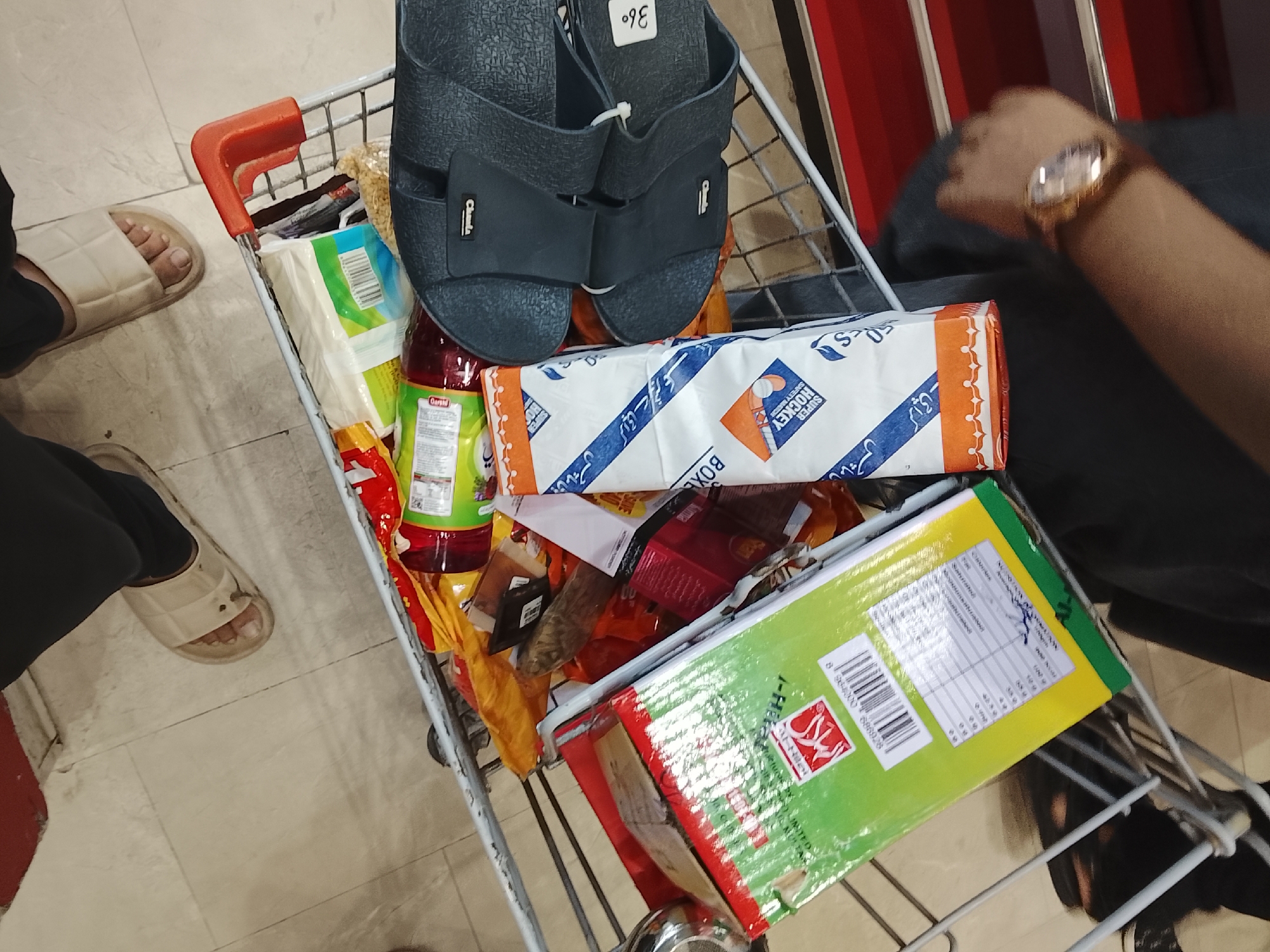 A shopping cart is filled with groceries and other items. The cart contains a box of Super Hockey safety matches, a bottle of Garshi brand food product, and a package of "Hira" brand goods. A pair of black sandals with the number "36°" are placed on top. The scene includes two individuals, one with a gold watch. The location appears to be a store in Gulgasht Colony, Pakistan, based on product information.