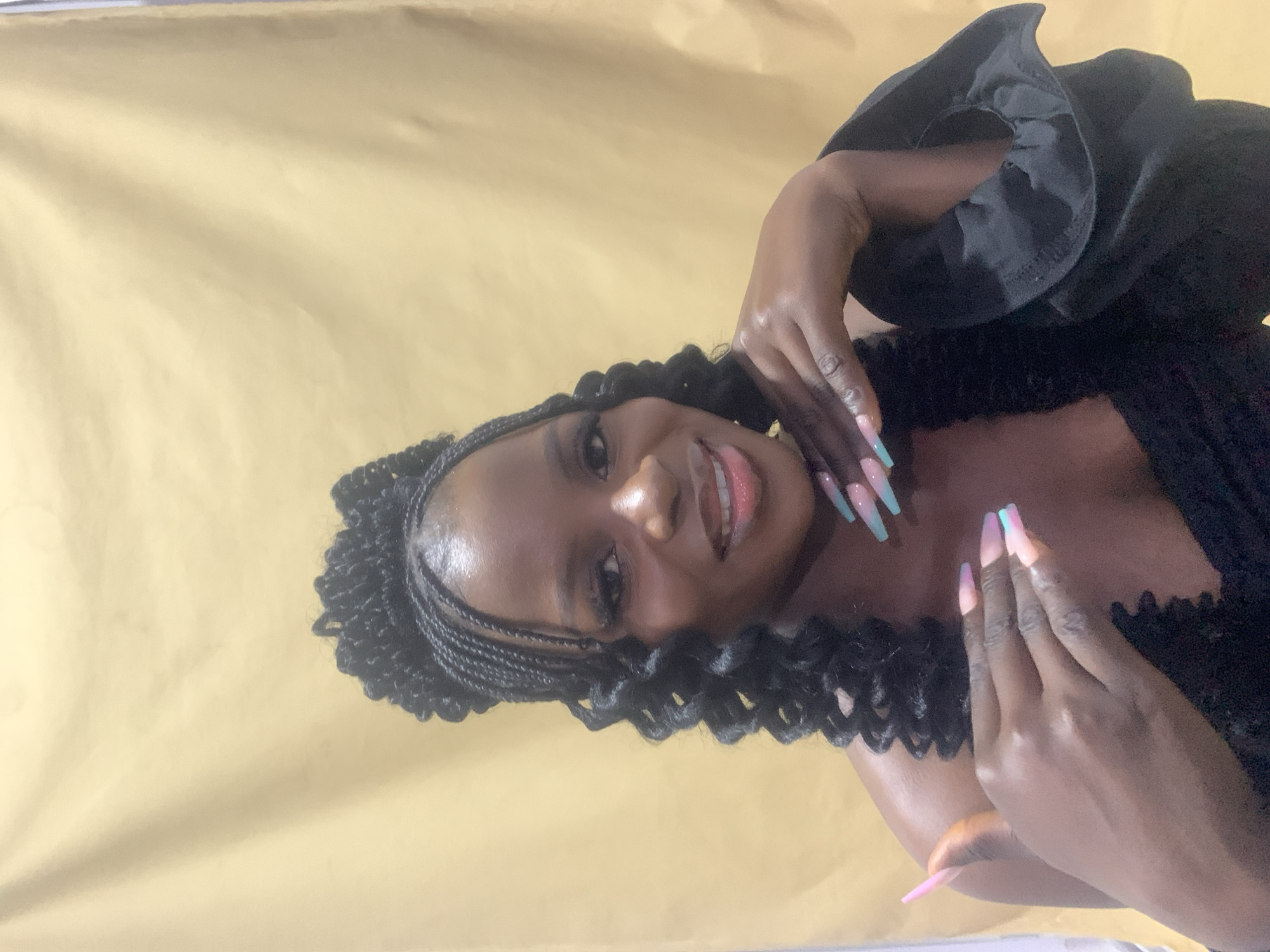 A woman with dark skin and black curly and braided hair is the main subject of this image. She is wearing a black outfit with a frilly or ruffled collar. Her nails are long and painted in pastel colors of pink, purple, and turquoise. She is smiling and her hands are near her face. The background is a solid, bright gold color. The image appears to be taken in a studio setting. The location in the city of Jos, Nigeria, cannot be verified from the image.