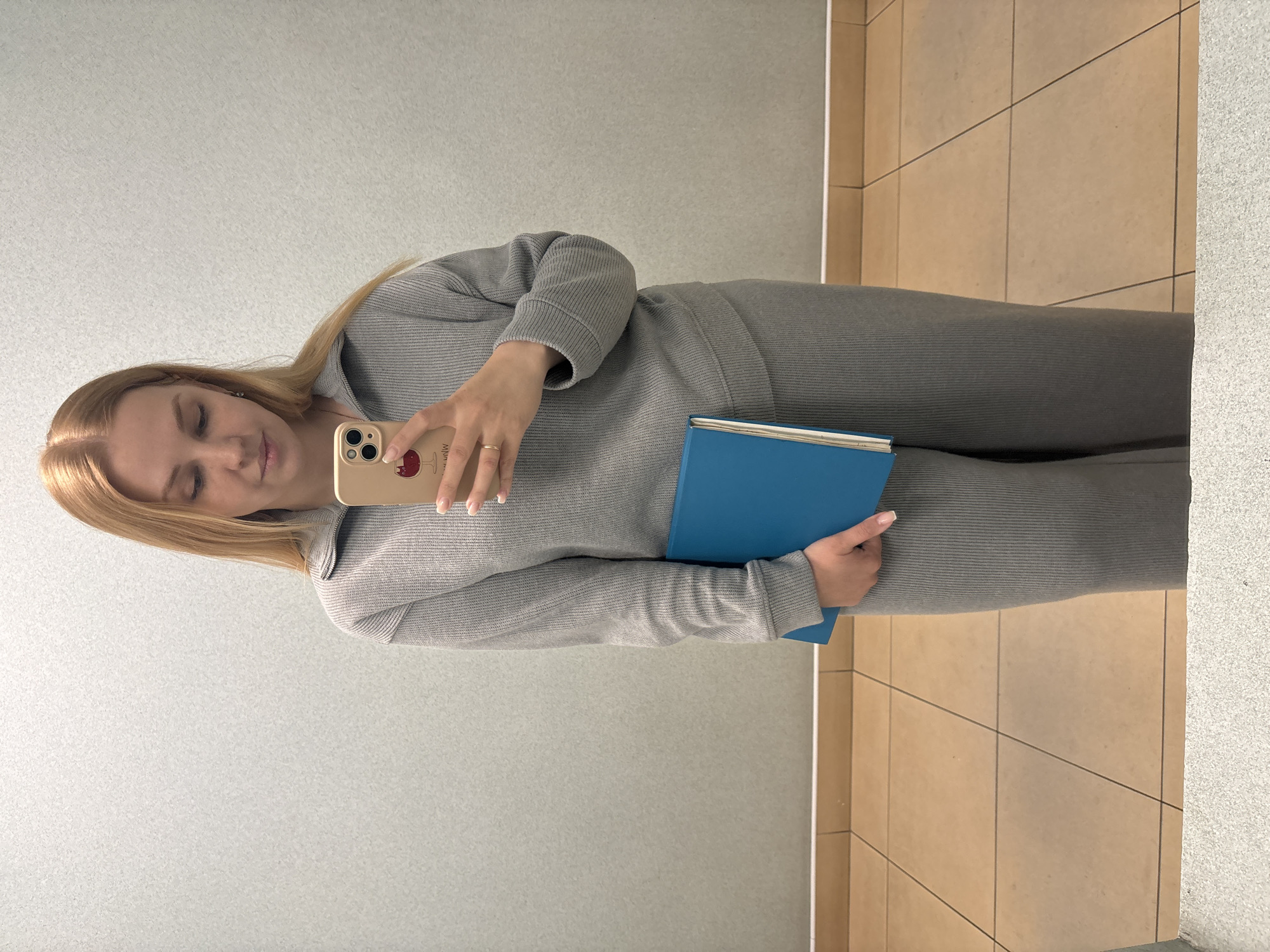 A person is taking a selfie in what appears to be a public restroom or bathroom. The person, presumably a woman, is wearing a gray ribbed-knit tracksuit. She is holding a blue book or folder in one hand and a smartphone in the other to capture the image. The background includes a gray tiled wall on the left and beige tiled wall on the right, suggesting a standard restroom design. No indication of Rusanovo, Russia can be found in the image.