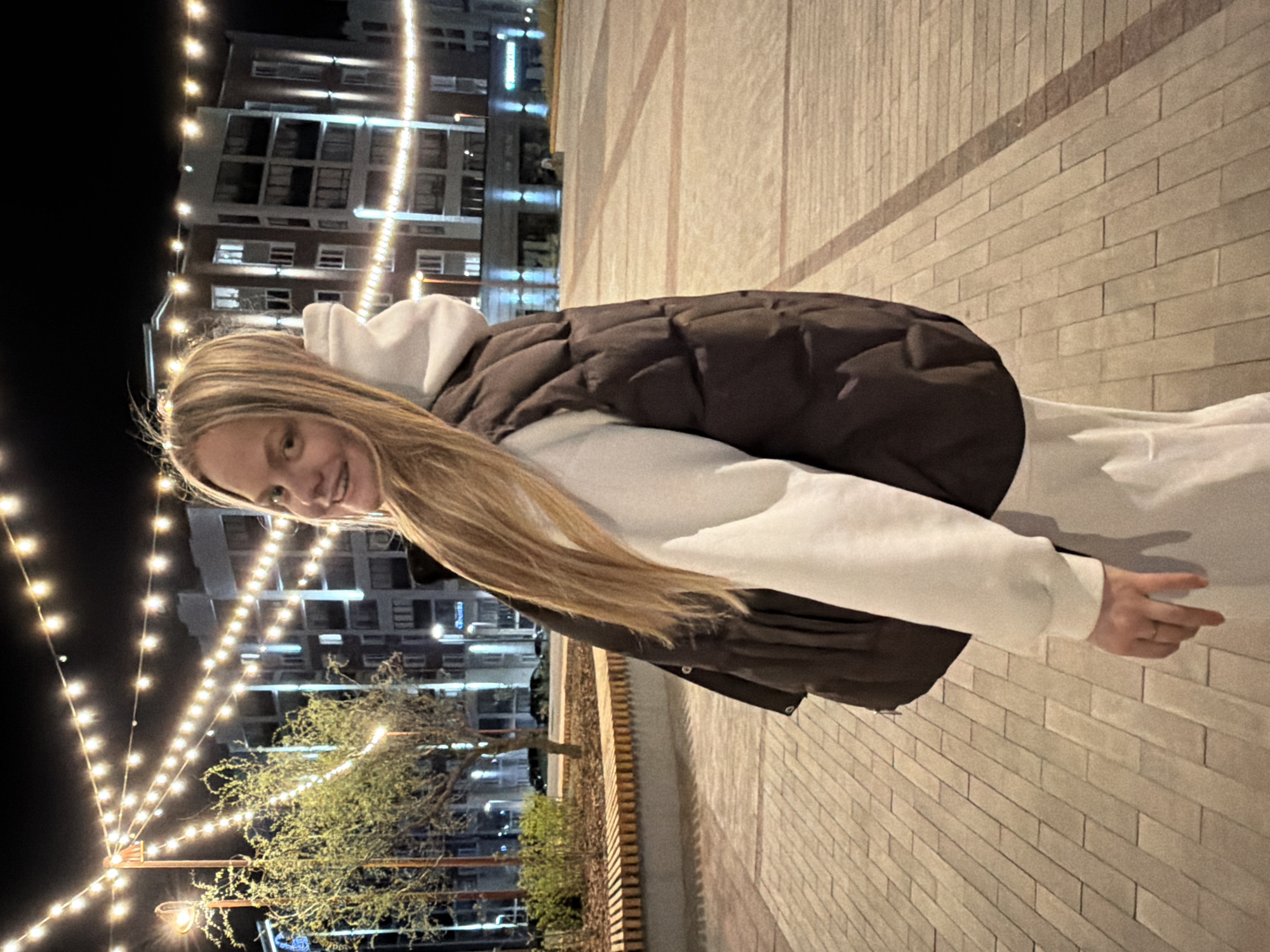 A woman is seen from a low angle, tilting her head to the side, possibly walking down a paved sidewalk in the city of Izhevsk, Russia, at night. She wears a white hooded sweatshirt with a dark, possibly black, puffer vest over it. Her long blonde hair is visible against the backdrop of a multi-story building illuminated with lights. The pavement is constructed of light-colored bricks, and a line of trees and bushes is visible along the sidewalk, illuminated with string lights. The background includes a dark night sky with a multi-story building featuring numerous windows, each emitting light.