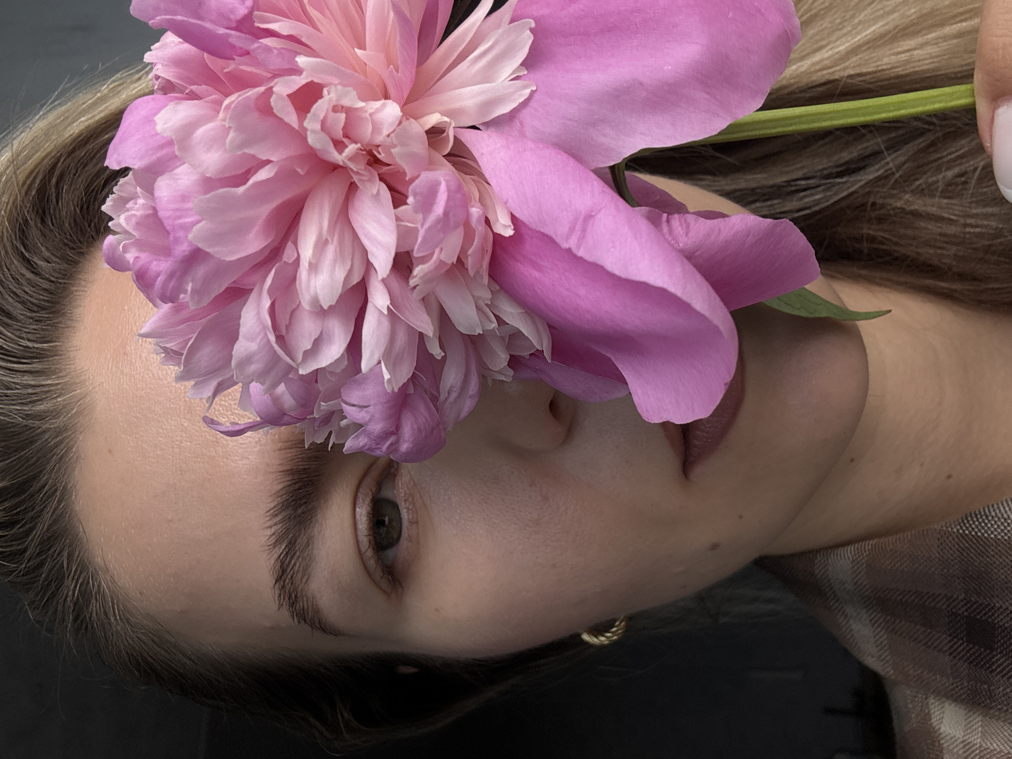 A close-up, vertical portrait captures a female subject, with the left side of her face mostly obscured by a large, vibrant pink peony. Her visible right eye, featuring a brownish-green iris, is directed straight at the viewer. Her light-colored hair frames her face, and her fair skin exhibits visible pores and a few small moles or freckles. She wears a small, gold-toned textured hoop earring in her visible ear. Her eyebrows are naturally full and well-groomed. Her lips are slightly parted, revealing a subtle lip color. She is dressed in a brown and tan plaid patterned garment, possibly a blazer or jacket. Her right hand, with short, light-colored manicured fingernails, gently grips the green stem of the peony at the bottom right of the frame. The peony itself displays numerous layers of soft, ruffled petals in various shades of pink, ranging from pale blush to a deeper fuchsia. The background is uniformly dark and indistinct, creating a strong contrast that highlights the subject and the flower. The location is Unknown, Unknown.