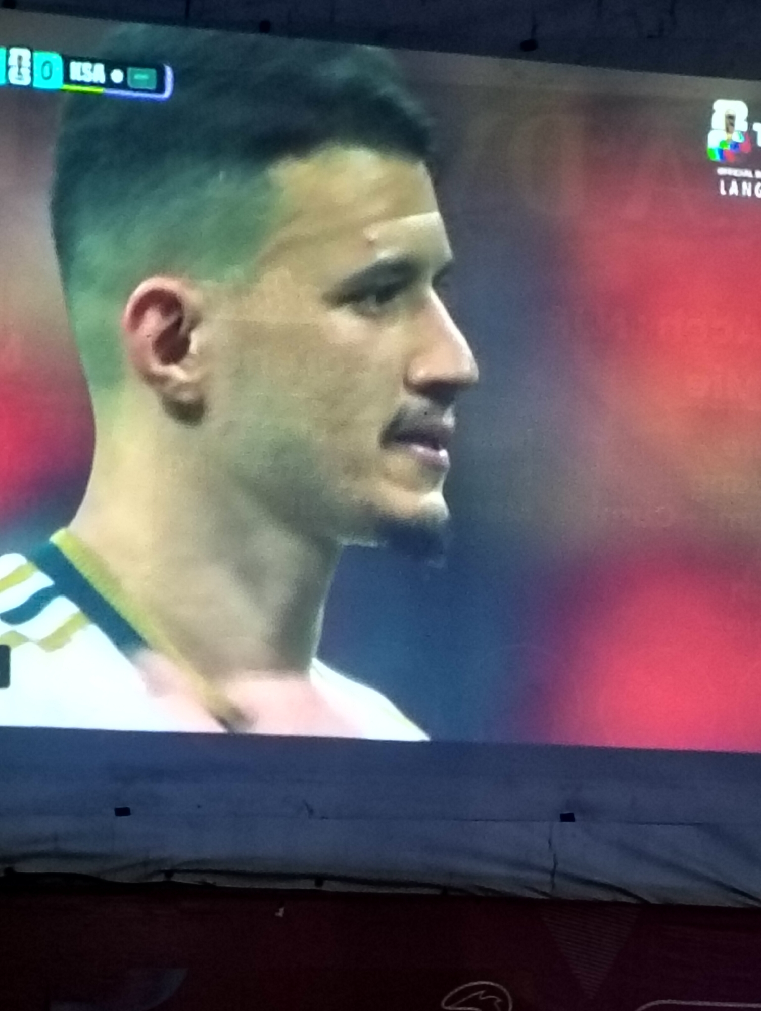 The image shows a close-up of a man's profile, likely a soccer player, displayed on a television screen. The man has dark hair styled in a fade, with a short beard and mustache. He is wearing a white jersey with a striped collar in yellow and dark green. In the top left corner of the screen, there is a scoreboard graphic showing "0" and "RSA" with a small green indicator. In the background on the right side of the screen, there are logos and text which are partially obscured and illegible due to image quality and glare. The television screen is situated in what appears to be a dimly lit indoor space, possibly a home or a gathering area, as evidenced by the dark horizontal band across the bottom of the image, which could be the top of another television or a piece of furniture. The overall lighting suggests it is nighttime or the room is dark. There is no visible indication of the city of Payagaboh, Indonesia within the image itself.