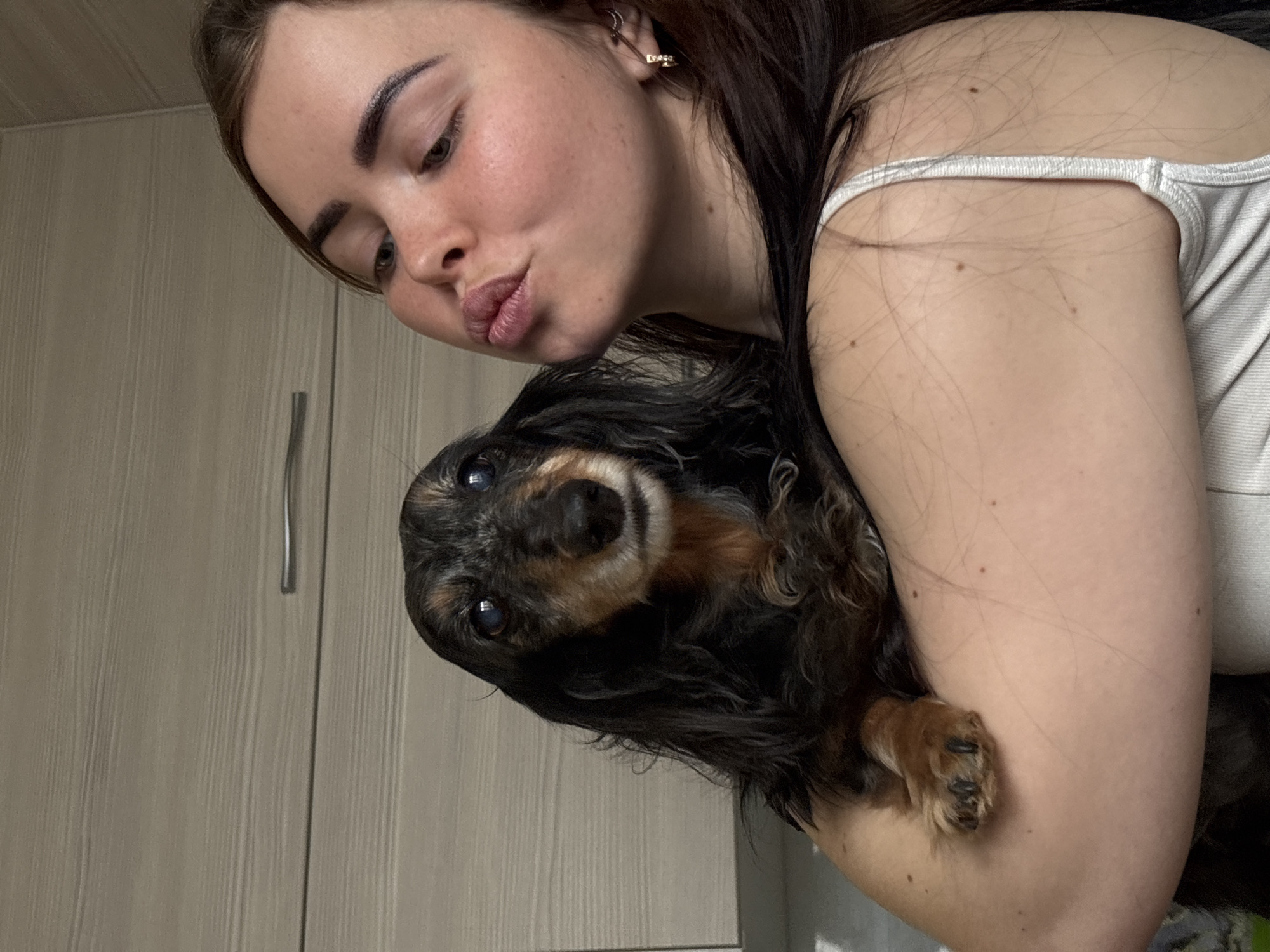 A person with long dark hair and fair skin is holding a black and brown dachshund dog. The person is wearing a white tank top and has an earring in their left ear. They are looking down with pursed lips. The dog is looking towards the camera with its paws resting on the person's arm. The background includes a light beige cabinet with a silver handle. It is not possible to determine the location in Izhevsk, Russia, from this image.