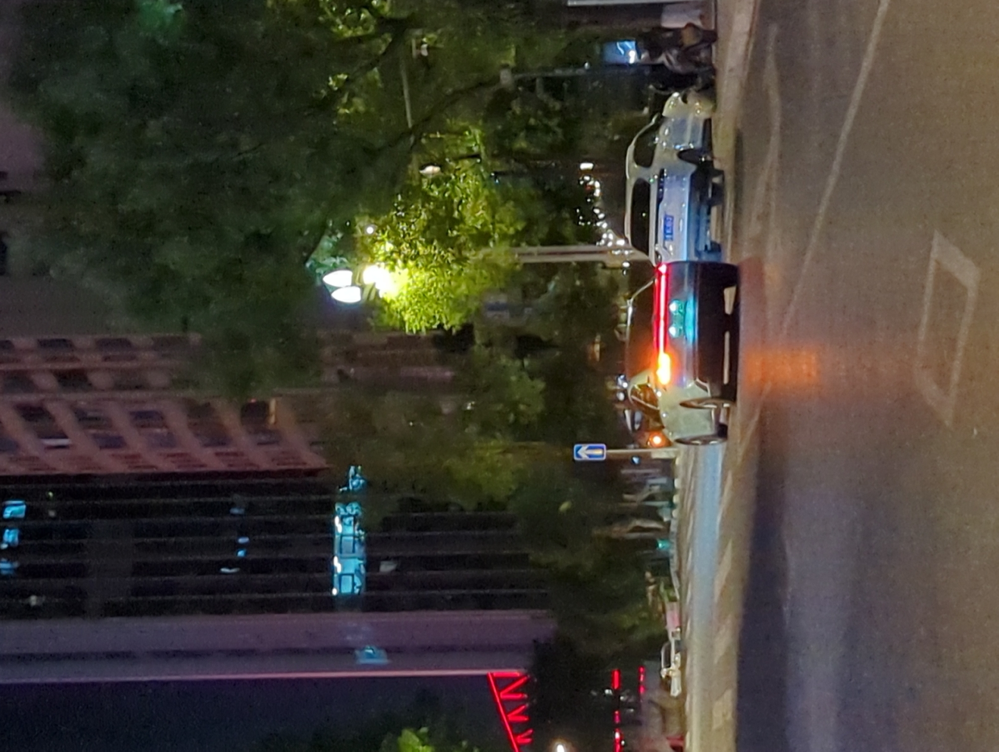 The image depicts a nighttime street scene in Jiangbeicheng Subdistrict, China. In the foreground, a road with a crosswalk is visible. Two cars are on the road; the car in the center is a light-colored SUV, and a darker sedan is to its left, its taillights illuminated. A motorcycle is parked on the right side of the road. In the background, a tall, modern building with many windows and lights can be seen, along with lush green trees and streetlights. The overall environment suggests an urban setting at night.