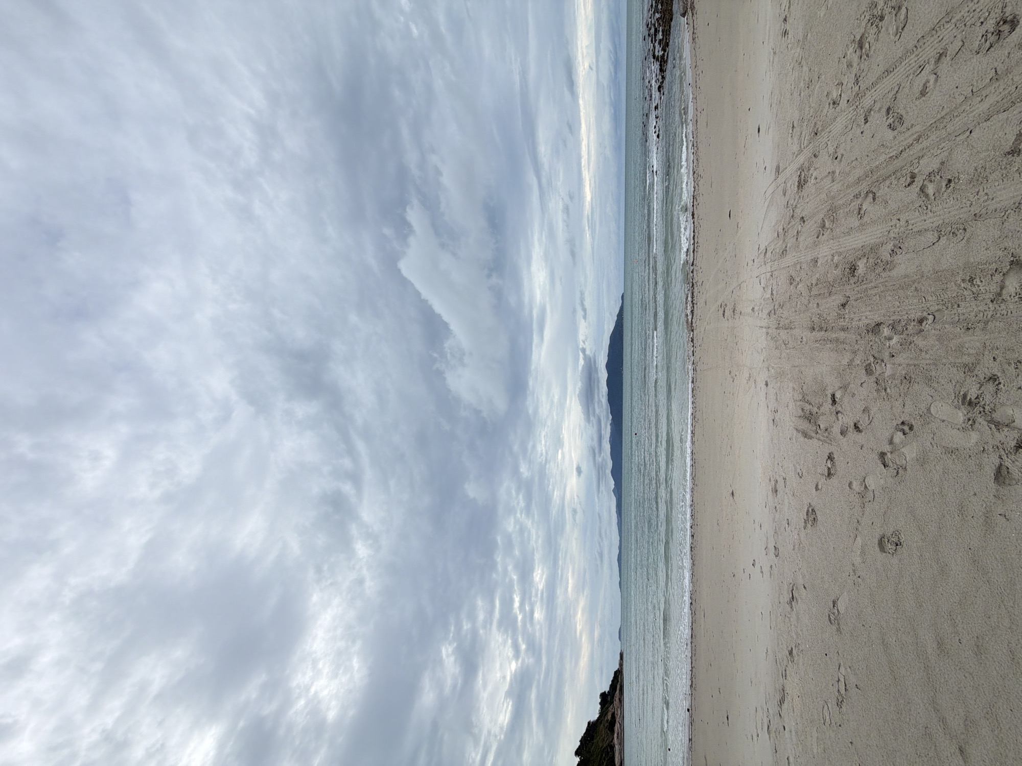 This media file depicts a tranquil beach scene under a cloudy sky. The orientation of the image is vertical. On the left side, a vast expanse of sky is filled with scattered, textured clouds, predominantly in shades of gray and white, suggesting an overcast day. The clouds are dense and appear to be moving. In the middle ground, a serene ocean meets the horizon, with the water appearing a muted blue-green. Distant, dark hills or mountains form a silhouette against the sky. The right side of the image showcases a sandy beach with subtle texture, marked by numerous footprints and tire tracks, indicating recent activity. The sand is light beige, and the waterline is visible with small waves lapping the shore. Towards the bottom left, a rocky, vegetated outcrop juts into the sea. The overall impression is one of a quiet, possibly cool day at a tropical beach. There is no visible text. The location is identified as Ko Samui District, Thailand.