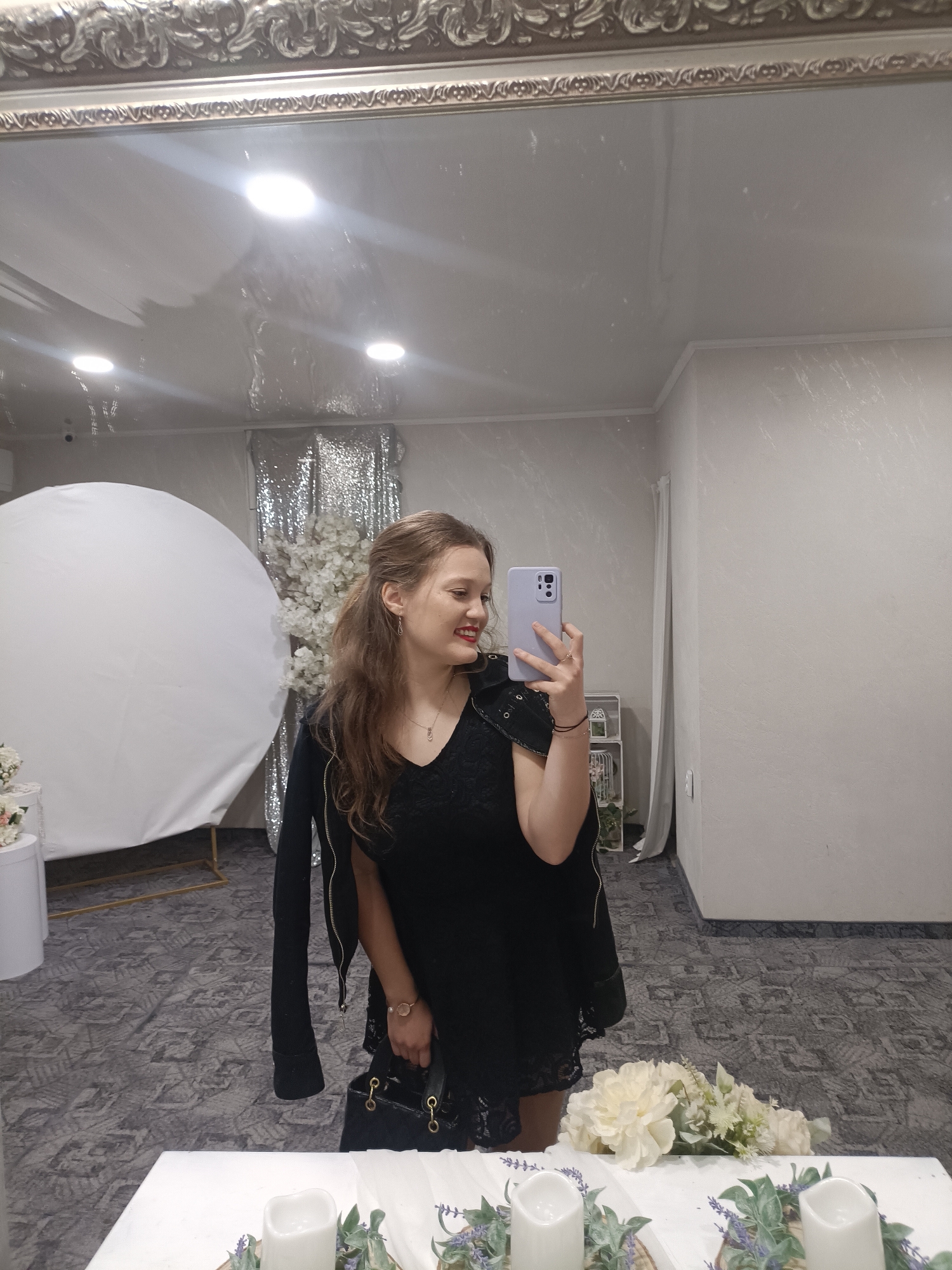 A fair-skinned woman with long brown hair partially pulled back is seen in a mirror selfie. She is wearing a black dress with lace trim and a black jacket draped over her shoulders. She holds a light purple smartphone in her right hand, obscuring part of her face. A black purse is in her left hand. She is smiling, with red lipstick visible.
The background includes a decorated event space. A large white circular screen is visible to the left, and behind the woman, there is a wall decorated with silver sequin fabric and artificial white flowers. Other decorations, including candles and flower arrangements, are on a table in the foreground.
The location is implied to be in Plovdiv, Bulgaria, based on user input but is not discernible from the image itself. The setting suggests an event such as a wedding or party. The flooring has a gray patterned carpet. The mirror itself has an ornate silver frame.