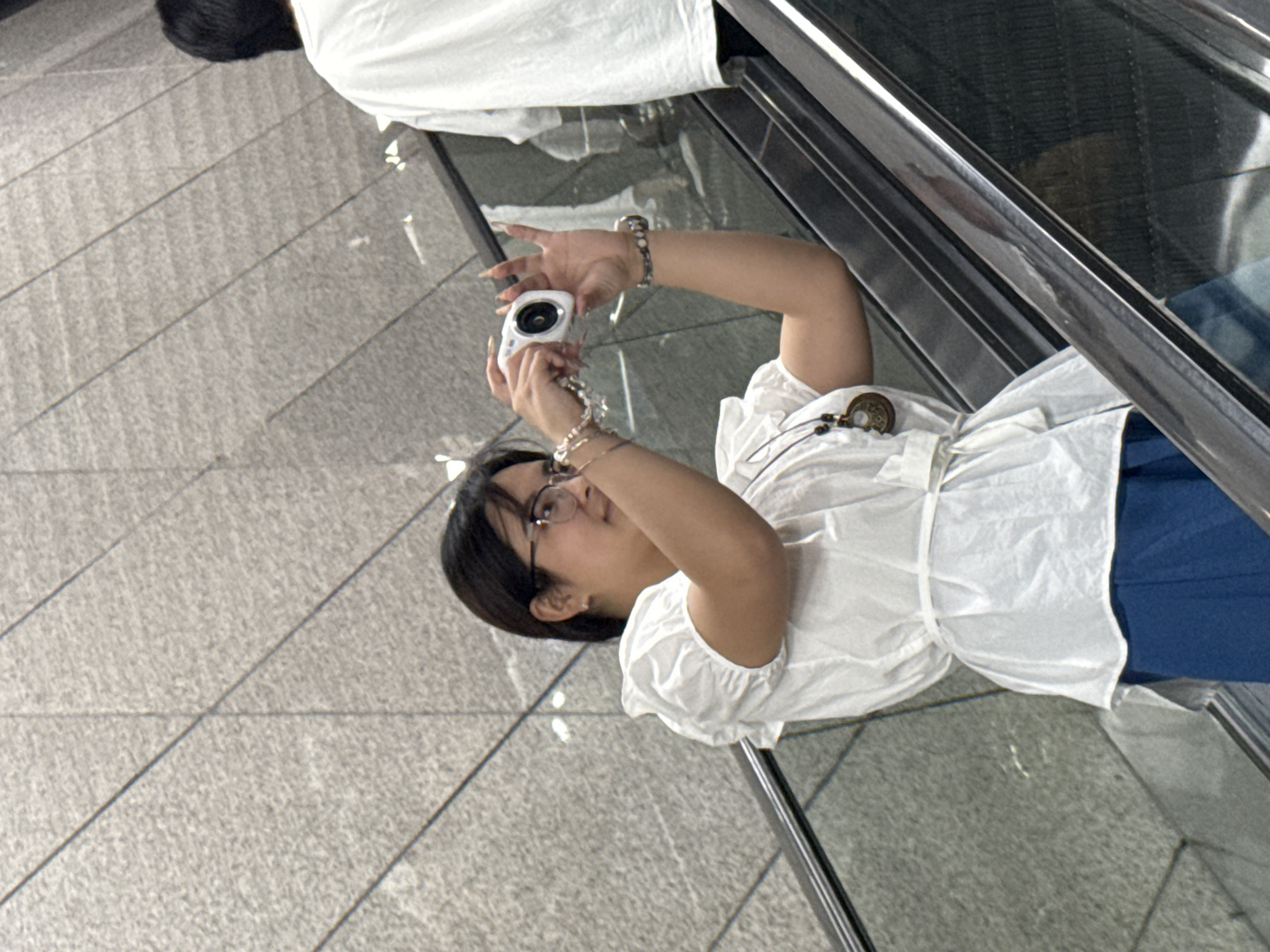 A person wearing glasses, a white top and blue pants is seen in an escalator holding a white camera taking a photo. An escalator and a tiled floor are visible. The location is Guang'an, China.