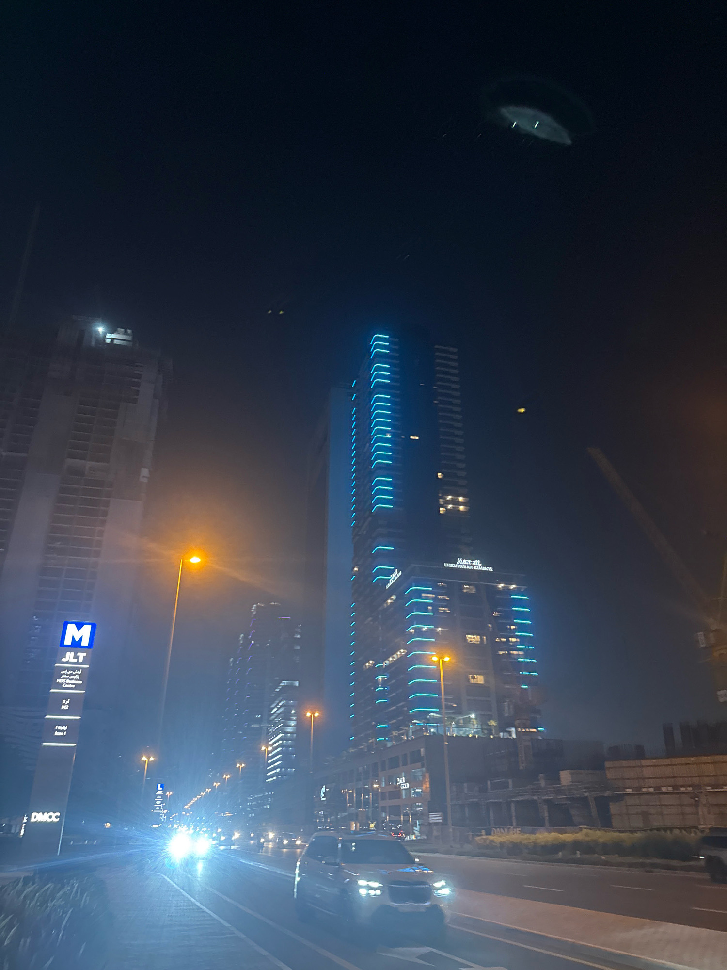 The image captures a nighttime street view in Dubai, United Arab Emirates, likely in the Jumeirah Lakes Towers (JLT) or Dubai Multi Commodities Centre (DMCC) area, as indicated by signage. Dominating the scene are modern skyscrapers, one of which is brightly illuminated with blue neon lights along its edges. Another tall building under construction is visible to the left, its skeletal structure illuminated by work lights. A street sign with the letter 'M' and "JLT" written on it is present on the left side of the road. The street itself is lit by streetlights, and the headlights of oncoming vehicles create a bright glare, suggesting a hazy or foggy atmosphere. Several cars are visible on the road, moving in both directions. The overall impression is of a bustling urban environment at night, with a focus on tall architecture and transportation.