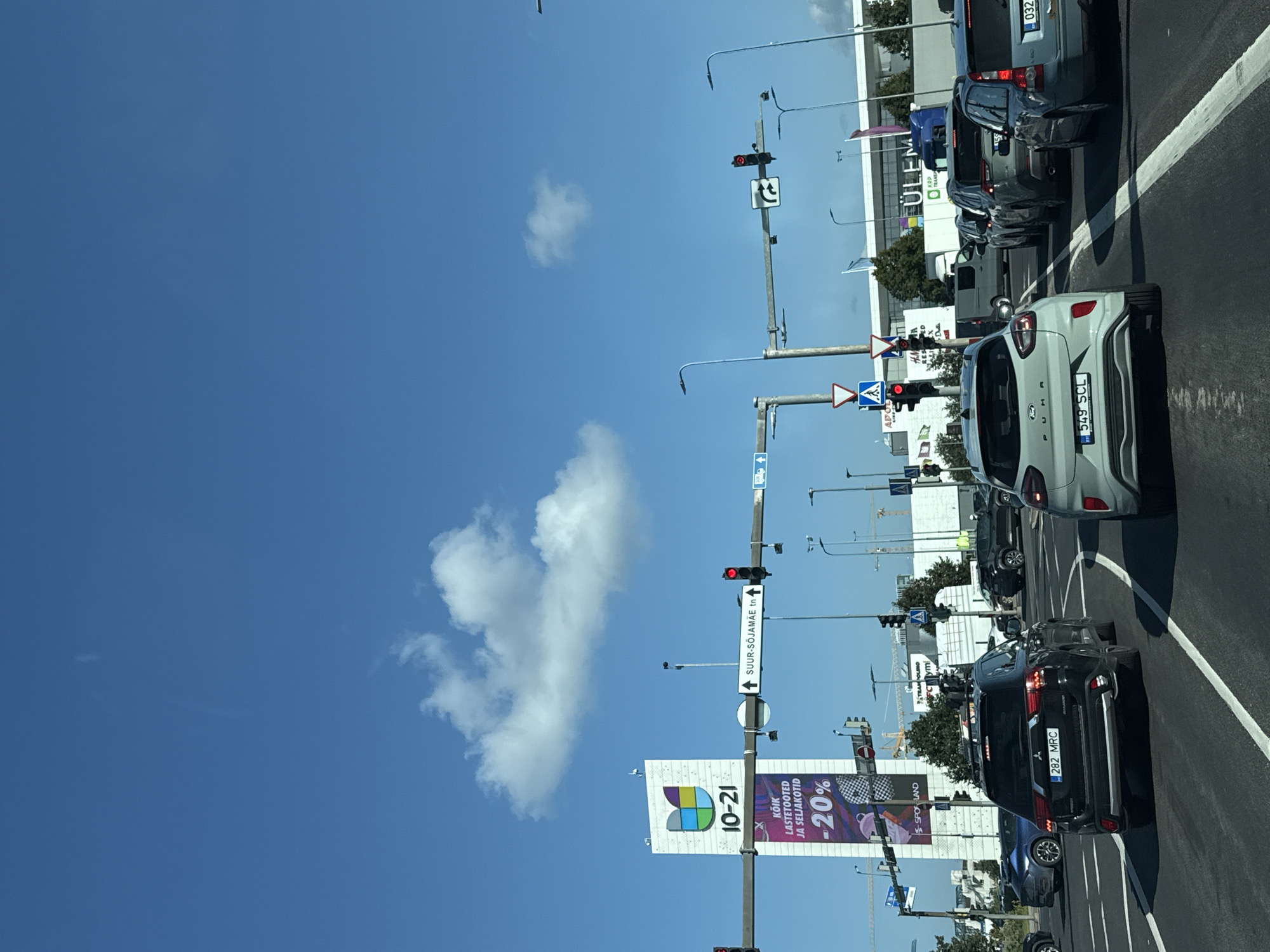 The image depicts a sunny day with a clear blue sky and scattered clouds. In the foreground, several cars are stopped at a traffic light intersection, indicating traffic congestion. The vehicles include a black SUV with license plate "282 MRC", a light-colored Ford hatchback with license plate "549 SCL", a dark blue station wagon, and other cars further back. Overhead traffic signals display red lights, stopping the vehicles.
A tall billboard on the left side of the image displays "10-21" and text in Estonian that translates to "Children's products and strollers -20%". This suggests a commercial area. Further in the background, a large building with the name "ÜLEMISTE" is visible, which is a well-known shopping center in Tallinn, Estonia. Other signs and streetlights are also present. The presence of traffic lights, multiple lanes of road, and commercial buildings points to an urban environment within Tallinn. The overall scene captures a moment of traffic flow in a commercial district of the city.