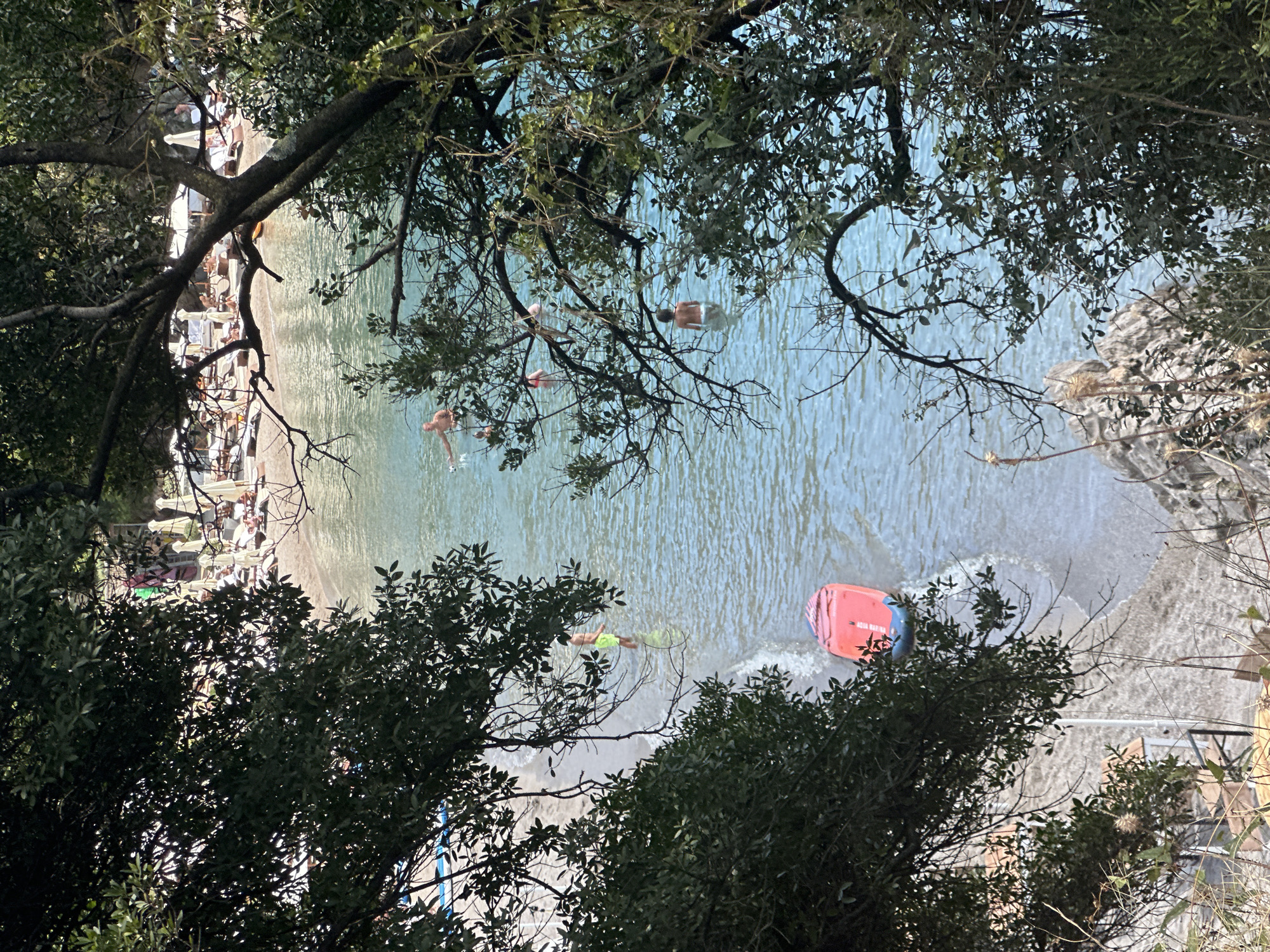 The image shows a view of a crowded beach from above, framed by lush green trees. The beach is lined with numerous lounge chairs and umbrellas, indicating a popular tourist spot. People are visible on the beach and in the clear blue water, some swimming and others relaxing. A red paddleboard is floating near the shore, and the gentle waves are lapping at the sandy beach. The overall scene depicts a vibrant summer day at a coastal resort. Based on the context of Budva, Montenegro, this scene is likely depicting Mogren Beach or a similar popular beach area within the city, known for its clear waters and scenic surroundings.