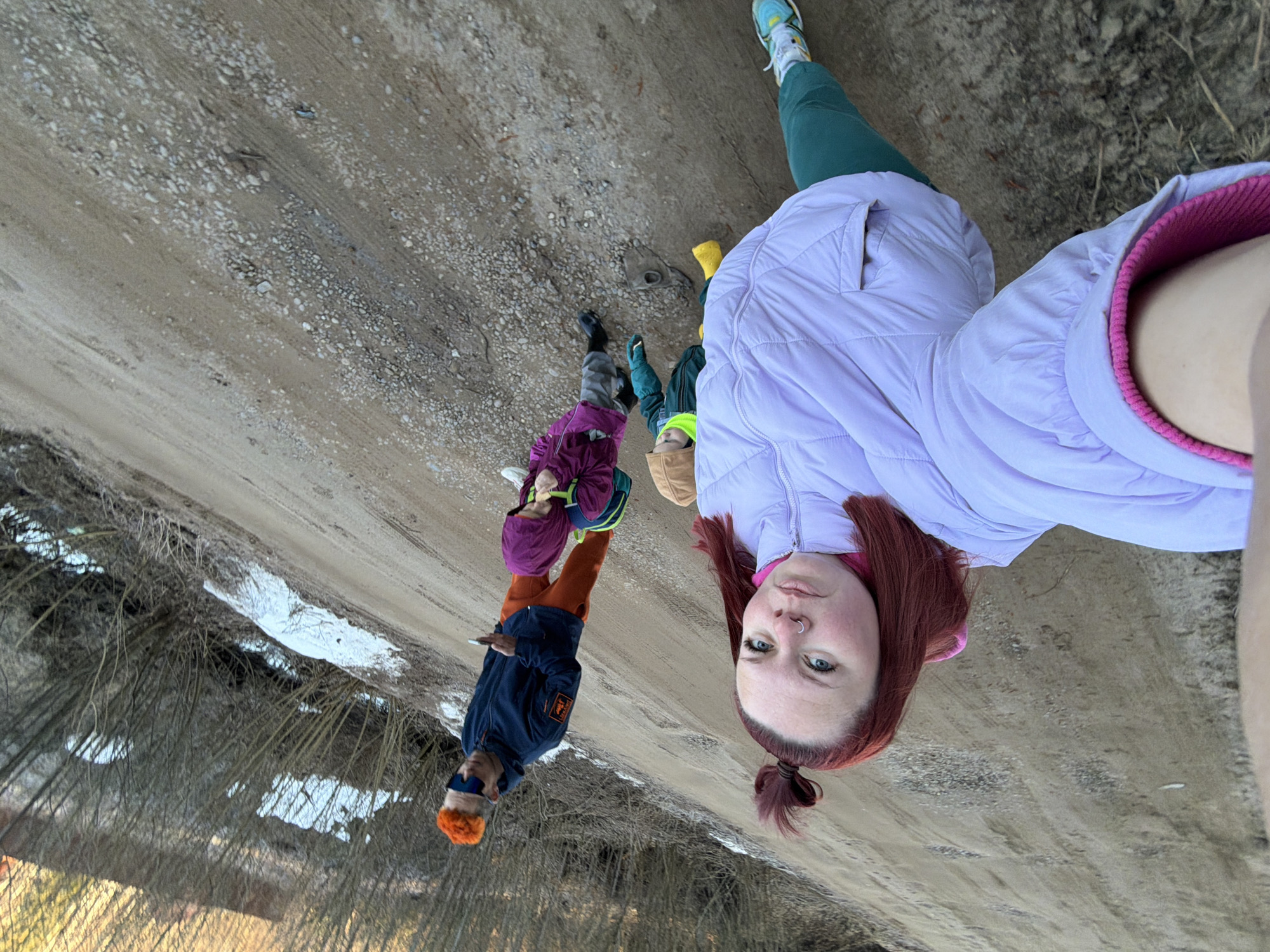 The image captures a scene on a dirt road or path on the outskirts of Chaikovsky, Russia, characterized by gravel and patches of melting snow. A person with reddish-auburn hair and a lilac-colored puffer jacket is positioned in the foreground, likely holding the camera for a selfie. Behind the person are several others, including at least two children wearing brightly colored jackets and hats. An adult male with bright orange hair is also visible. The group appears to be standing or walking together along the path. Patches of dried vegetation and bare trees are visible along the side of the road, indicating an early spring or late autumn season.