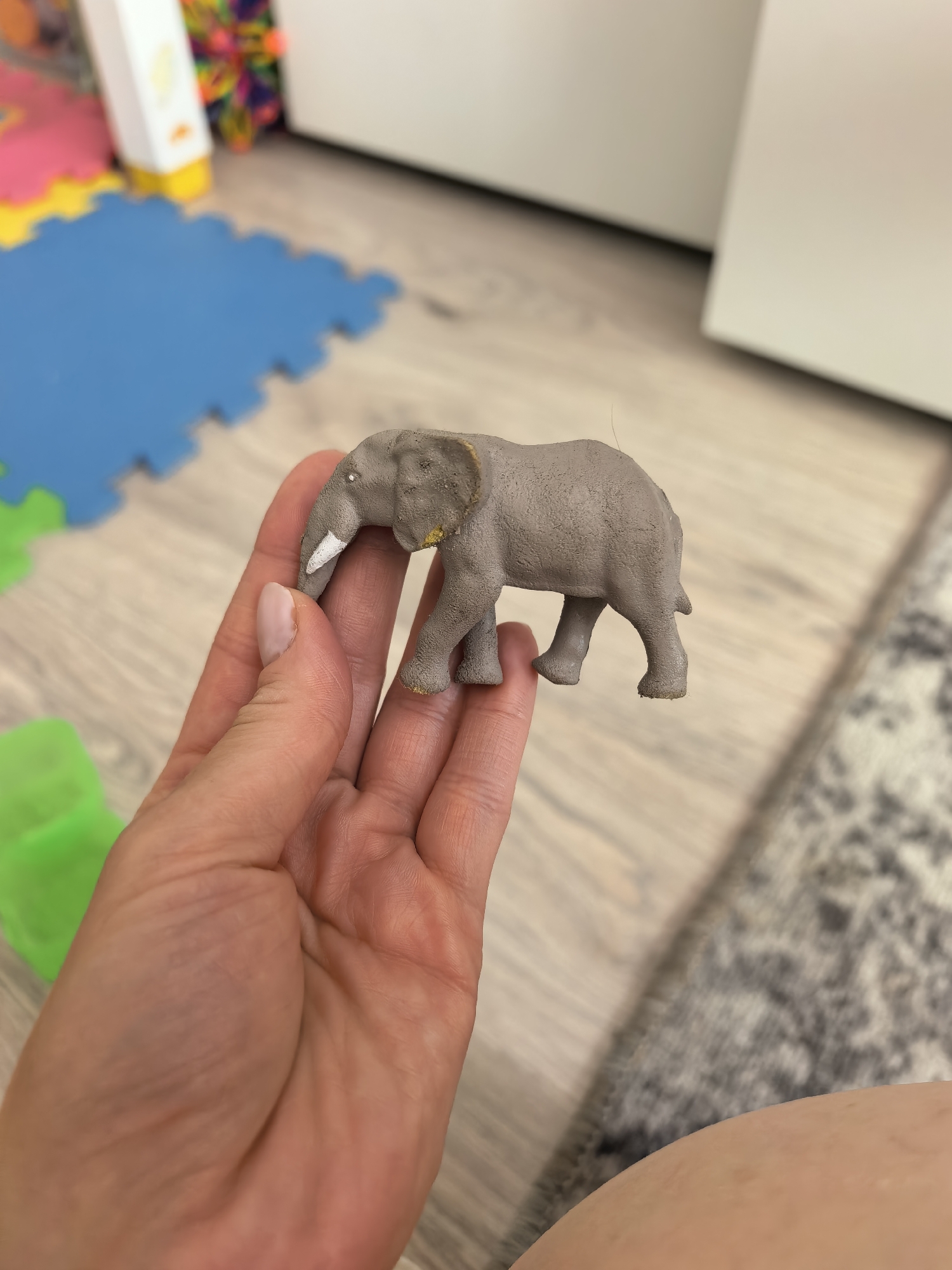 A person is holding a gray toy elephant in their hand. The elephant has white tusks and some gold paint on its ear. The background includes children's play mats, a toy, a cabinet, and a rug. There is no indication this photo was taken in Sterlitamak, Russia.