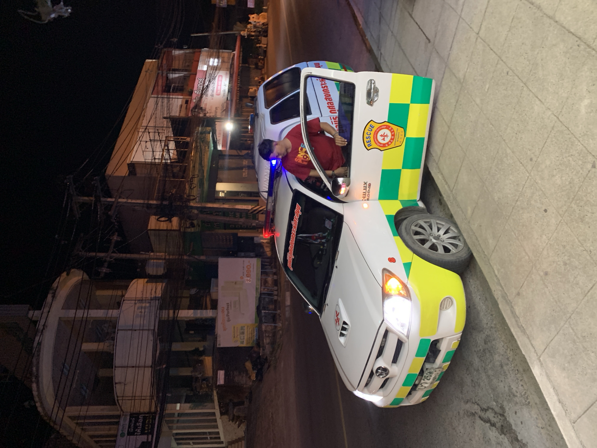 In Baan Bo Phut, Thailand, a white ambulance with green and yellow checkered markings is parked on the side of a road at night. A young man wearing a red t-shirt stands in the open driver's side door, leaning against the vehicle. The ambulance has flashing red and blue lights on its roof. Behind the ambulance, several buildings are visible, some illuminated and others in darkness, with overhead power lines strung across the street. A sidewalk with concrete paving stones is in the foreground.