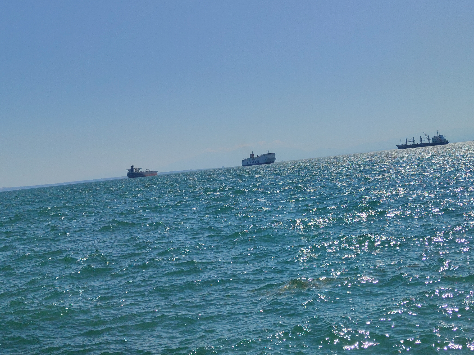 A wide shot captures the Aegean Sea under a clear, pale blue sky. The water is a deep teal, rippled and sparkling with sunlight. In the distance, three large cargo ships are visible on the horizon, spaced out across the frame. The ship on the left is a dark-hulled tanker. The middle ship is a white passenger ferry, identifiable by its superstructure and the text "RIO KINALIS" on its side. The ship on the far right is a dark cargo vessel with cranes. The coastline is faintly visible as a hazy line in the far distance. The scene appears to be taken from a boat or coastal vantage point within or near the Thessaloniki Municipal Unit, Greece, given the presence of these large vessels in a body of water characteristic of the region.