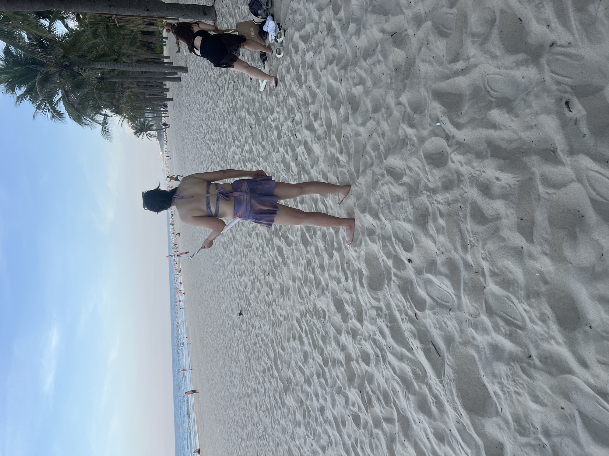 The image depicts a beach scene in Đà Nẵng, Vietnam, under a bright, clear sky, suggesting it is daytime, likely late morning or afternoon given the quality of light. The foreground is dominated by the textured sand of the beach, with visible footprints and undulations.
In the middle ground, two individuals are the primary subjects. One person, seen from the back, is wearing a purple-toned bikini and skirt, with their back to the camera, facing the ocean. They appear to be holding a selfie stick or a similar device. To their left and slightly further back, another person is lying down on the sand, wearing a black outfit.
The background reveals a bustling beach environment. A line of palm trees stretches along the edge of the beach, indicating a tropical or subtropical location. Beyond the palm trees, several buildings are visible, hinting at a developed coastal area. The ocean is present, with many people scattered along the shore and in the water, suggesting a popular beach destination. Red flags can be seen in the water, possibly marking swimming zones or areas with specific conditions. The overall atmosphere appears relaxed and lively, characteristic of a sunny day at the beach.