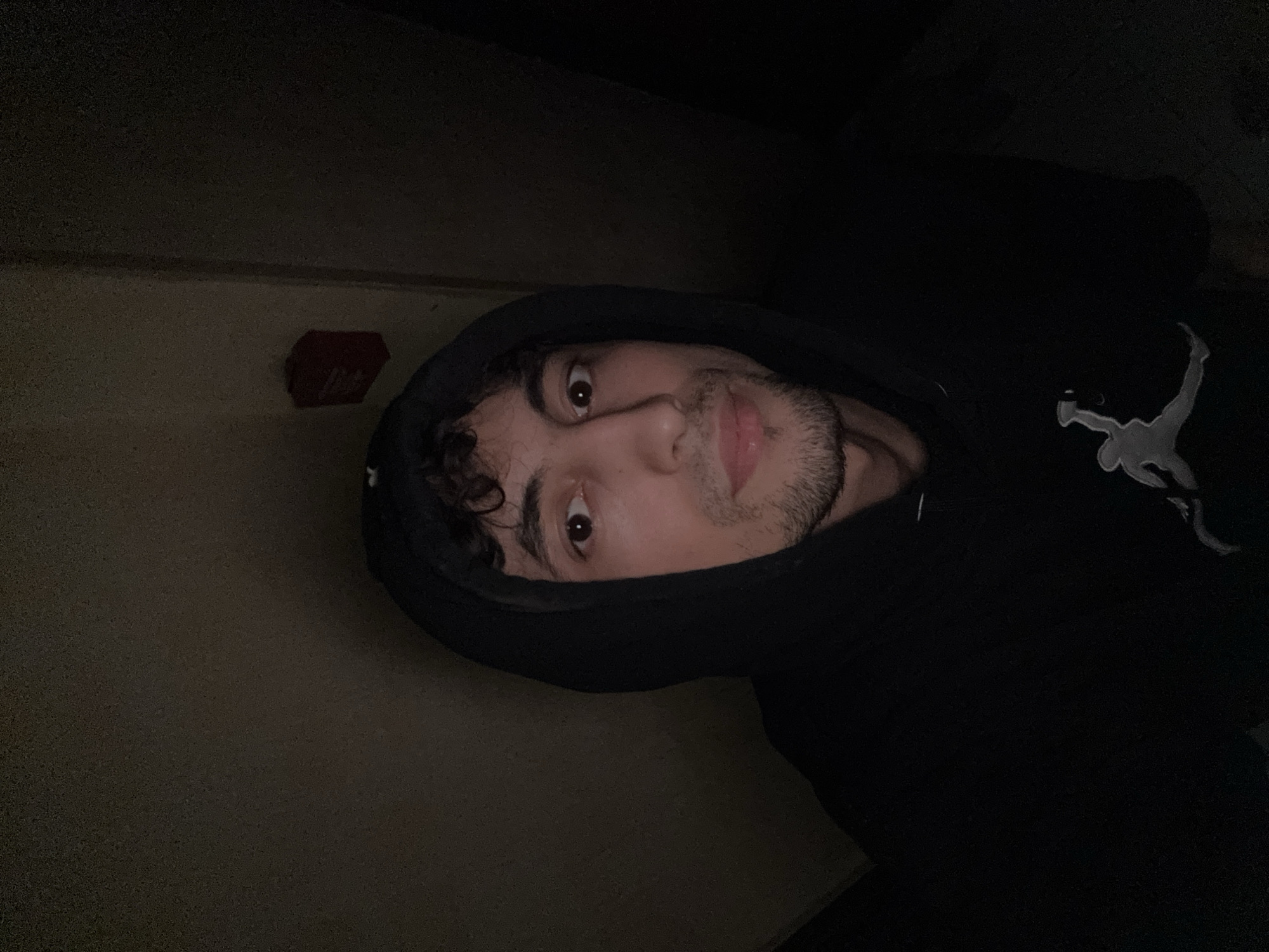 This media file is a close-up, low-angle selfie of a young man wearing a black hoodie with the hood pulled up. The man has curly dark hair, brown eyes, and a light beard. He is looking directly at the camera with a neutral expression.
The background is dark and appears to be indoors, possibly a hallway or room with minimal lighting. To the left of the man, a small red object, possibly a fire alarm or emergency button, is visible on a plain wall. The lighting is dim, casting shadows and highlighting the man's face. The overall mood is somewhat mysterious due to the low light and the hoodie obscuring most of his features. There is a faint silhouette of a logo on the hoodie, resembling the Jumpman logo associated with Nike. No time of day or weather cues are apparent.