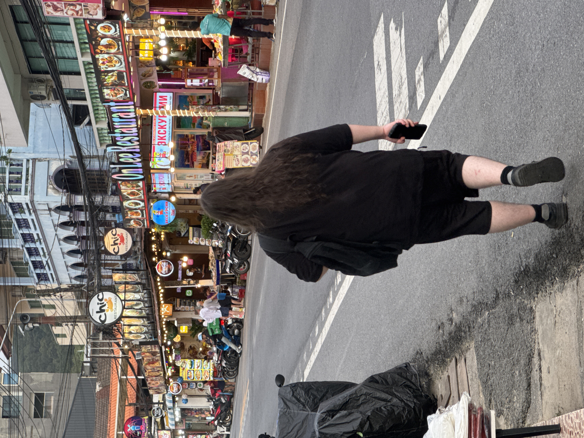 In Karon, Thailand, a person with long, dark, wavy hair walks away from the camera down a city street. They are wearing a black t-shirt and black shorts, with dark socks and shoes. The person is holding a dark-colored smartphone in their right hand. The street is paved and has a white pedestrian crossing marked on it. To the left of the street are several parked motorcycles, some covered with black tarps. A stall with stacks of folded fabric is also visible on the left. On the right side of the street, a row of restaurants and shops line the sidewalk. Several signs are visible, including "chic RESTAURANT" and "On Lee Restaurant". Lights are illuminated on the signs and storefronts. People are visible inside and outside some of the establishments, including individuals working at food stalls. Buildings rise up on either side of the street, with visible electrical wires strung overhead. The overall scene depicts a bustling commercial street.
