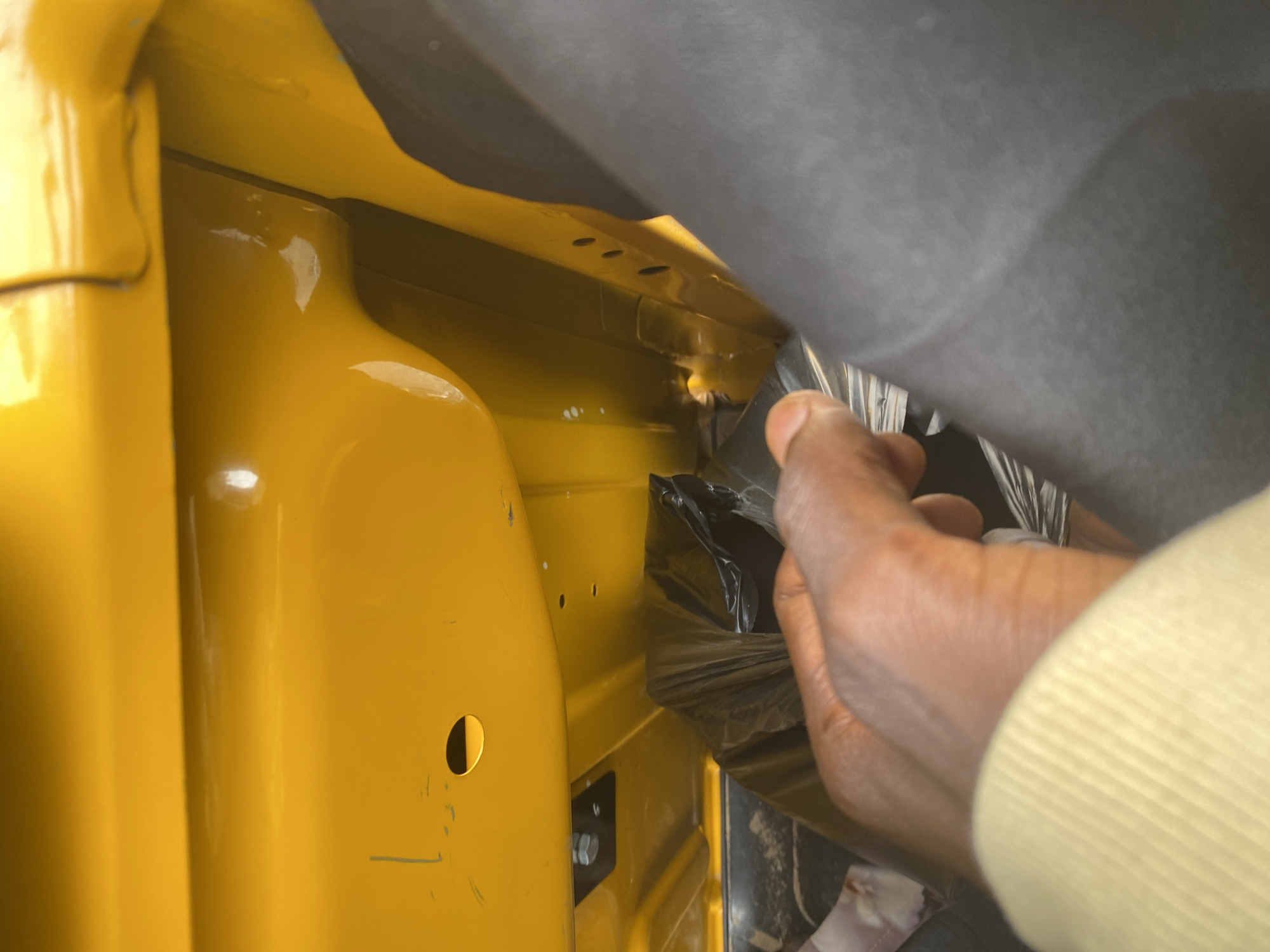 The image shows a close-up of a yellow metal surface, likely the interior of a vehicle or industrial equipment. A person's hand, with dark skin and a light-colored sleeve, is reaching into a black plastic bag that is tucked into a crevice of the yellow surface. The yellow surface has several small holes and a larger circular opening, possibly for bolts or fasteners. There is a bolt with a black bracket attached to the surface. The background is dark and out of focus, with a hint of dark fabric on the right side of the image, suggesting the presence of a person. The lighting suggests an indoor or shaded environment. The overall scene is devoid of any clear context related to Jalingo, Nigeria, as there are no identifiable landmarks or contextual clues.