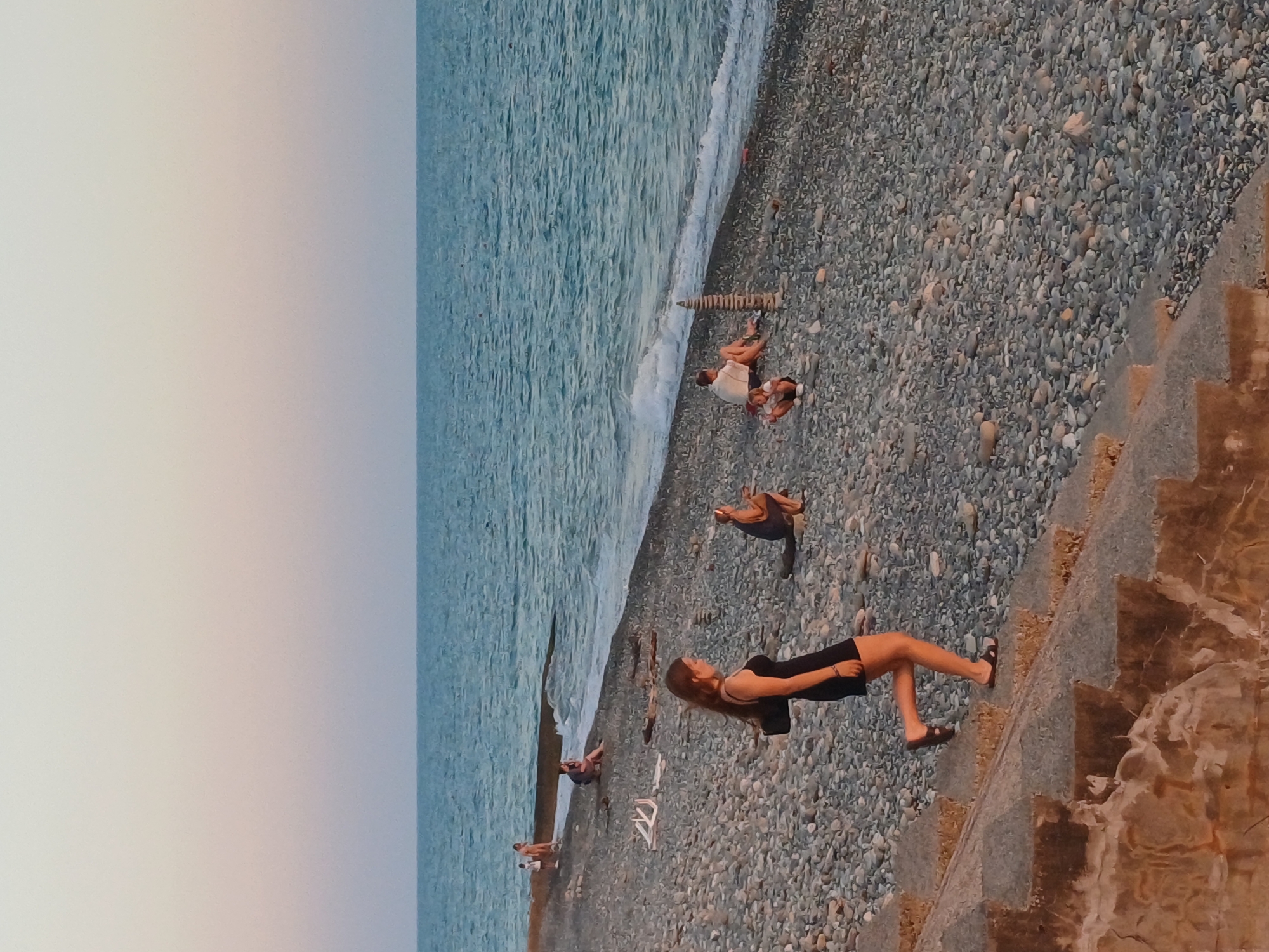 This photograph depicts a pebble beach in Sochi, Russia, during the golden hour. The sky is a gradient of pale pink and blue, meeting the turquoise sea. Several individuals are scattered across the beach, engaged in various activities. In the foreground, a young woman in a black dress walks down stone steps, her left leg forward, wearing sandals. To her right and further down the beach, another woman crouches near a pile of stacked stones. In the midground, a person sits on the beach, facing away from the camera, while a child squats near them. Further out, two individuals are seated near the water's edge. In the background, a concrete breakwater extends into the sea, and a few people are visible near it. The beach itself is covered in smooth, grey pebbles of varying sizes.