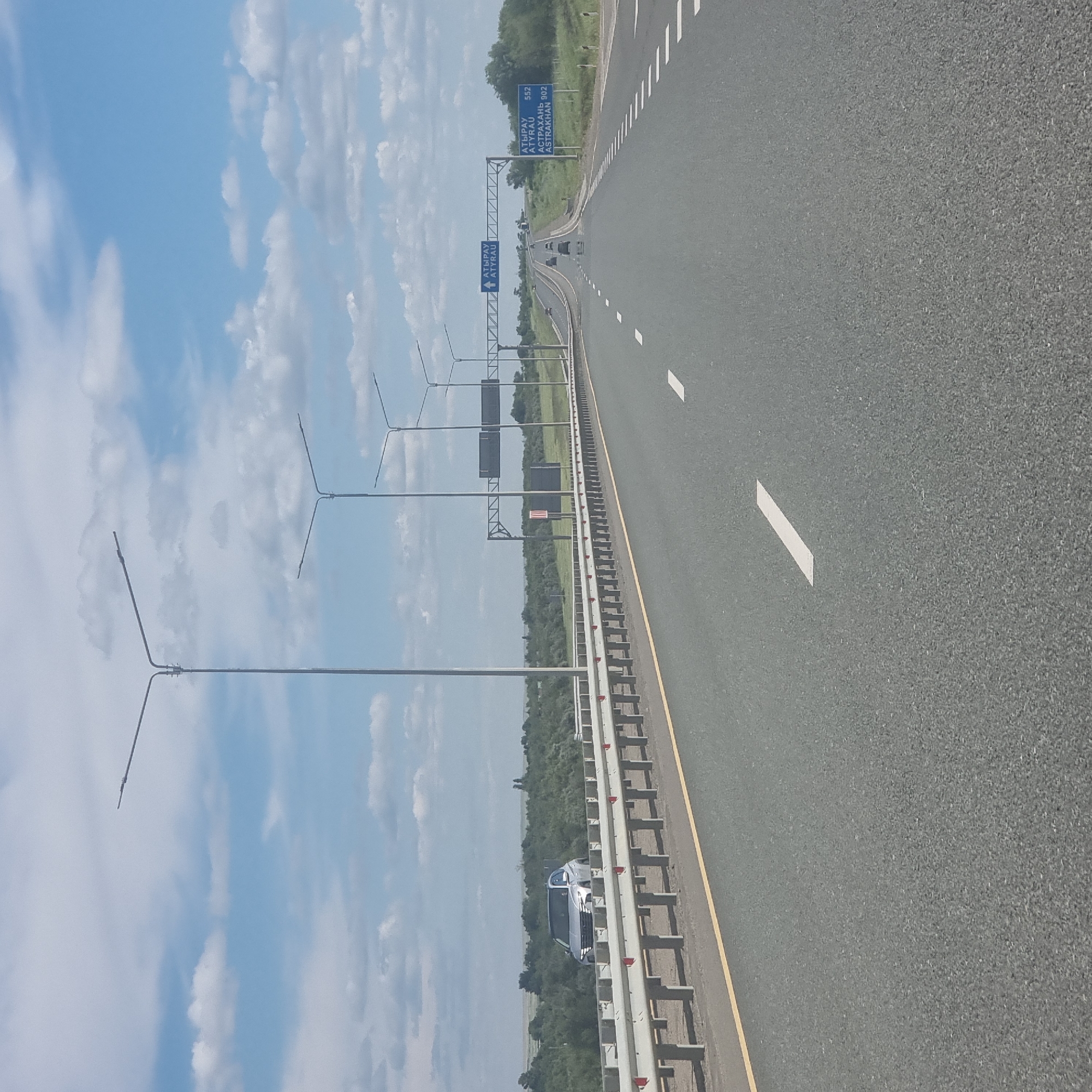 The image shows a highway leading towards a distant horizon under a partly cloudy sky. Tall lampposts line the right side of the road. A silver SUV is visible in the distance on the left side of the road, behind a guardrail. A blue road sign is visible on the right side of the road indicating directions and distances to Atyrau (552 km) and Astrakhan (902 km). The road is asphalt with clear lane markings. The environment appears rural with vegetation and trees visible in the background. The location is identified as the highway near Alğa, Kazakhstan.