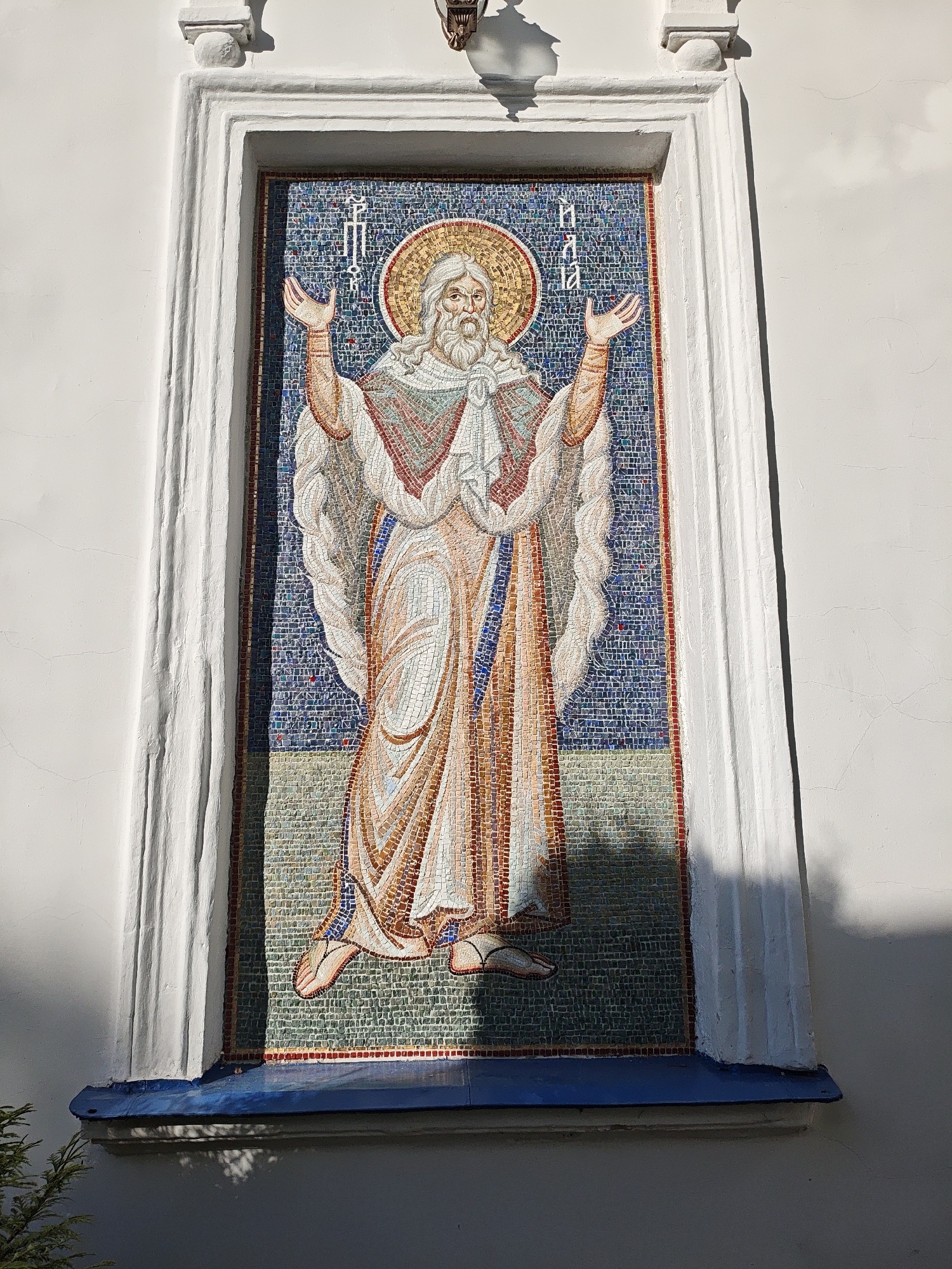 The image shows a mosaic depicting a standing figure of a bearded man with a halo, possibly a religious figure. The mosaic is set in a white framed niche on a light-colored building exterior. The background of the mosaic is a dense pattern of blue and white tiles. The mosaic itself features earth-toned robes and white hair. Above the figure are Cyrillic characters. The building's exterior is plain and white with simple architectural details. The base of the niche is painted blue. The location is Мамонтово, Russia.