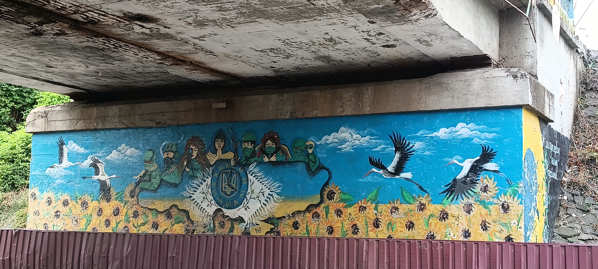 The image shows a mural painted on the concrete wall beneath a bridge. The mural depicts a row of figures wearing green military-style uniforms and face coverings, with some wearing helmets. In the center of the mural is a blue and yellow emblem with a clock face and what appears to be a trident symbol. The bottom of the mural features a field of sunflowers under a blue sky with white clouds. Two storks are painted in flight over the sunflowers. The mural is located on a wall in Kamianka, Ukraine, as indicated by the presence of Ukrainian national symbols and motifs. The bridge structure above the mural is made of concrete and shows signs of wear and tear. A brown corrugated fence runs along the bottom of the image, obscuring the base of the mural.