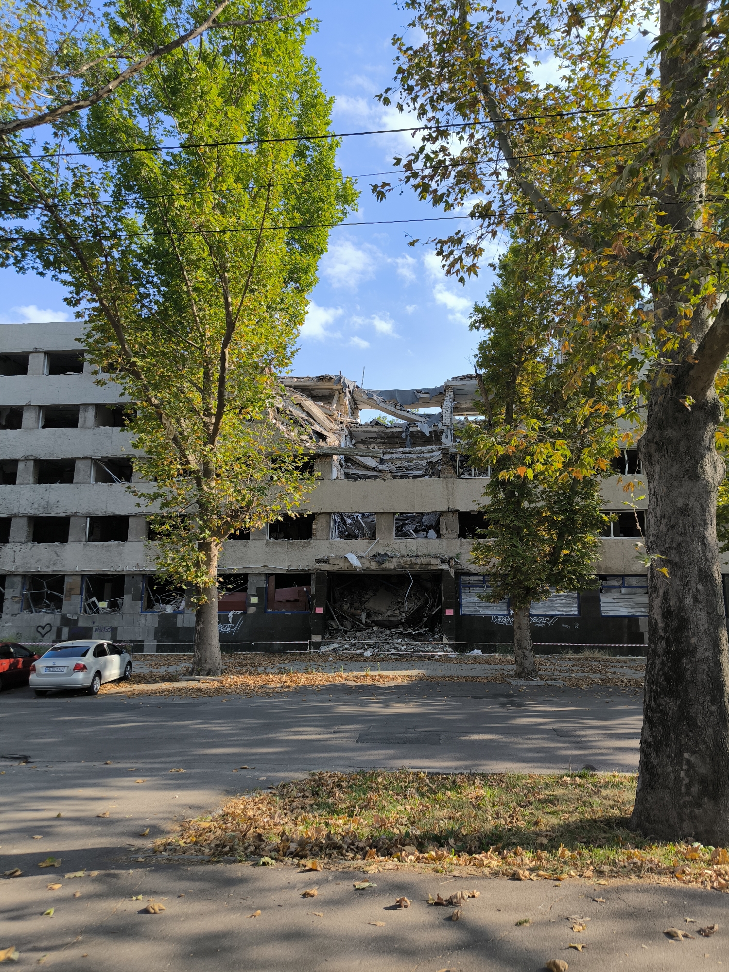 A damaged apartment building stands in Mykolaiv, Ukraine, under a partly cloudy blue sky. The building, appearing to be at least four stories tall, has suffered significant structural damage, with its roof and upper floors collapsed and exposed. The facade shows numerous empty window frames, and debris is visible at the base of the structure. A white car is parked in front of the building on the asphalt road, with fallen autumn leaves scattered on the ground. Two large trees, one with vibrant green leaves and the other with leaves showing autumn colors, flank the building, partially obscuring the view of its damaged sections. Power lines are visible running between the trees.