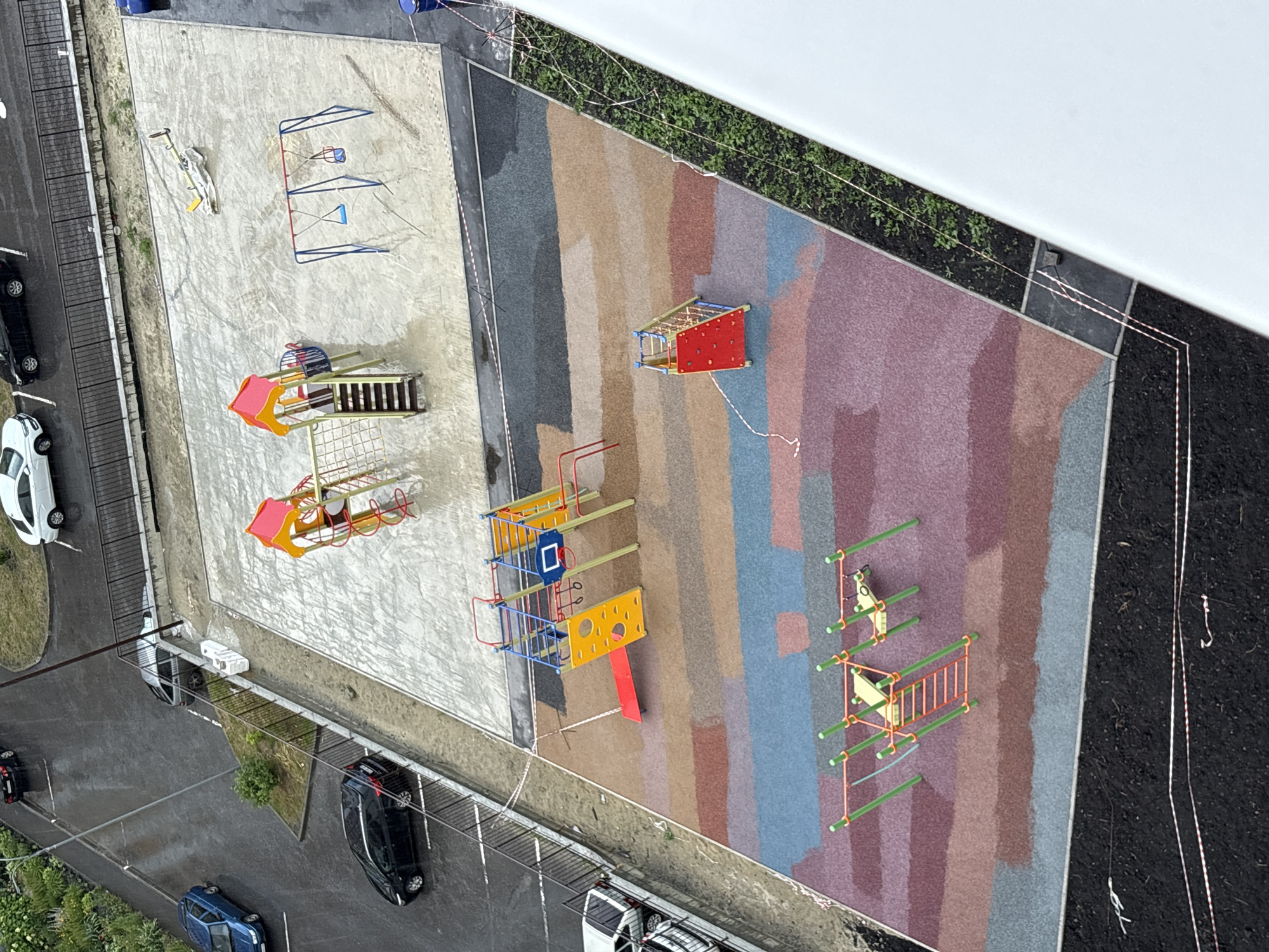 The image shows an overhead view of a newly constructed playground in the city of Засечное, Russia. The playground is situated on a multi-colored, impact-absorbing surface with sections of gray concrete. To the left of the playground, a paved road with painted parking spaces is visible, with several cars parked, including a blue hatchback and a black sedan. Rain is falling, and the road and playground surfaces are wet.
The playground equipment includes a climbing structure with a red slide, a swing set with two blue swings, and a separate, smaller climbing structure with a red base and netting. Further to the right, another section of the playground features a climbing frame with green poles and a red ladder, and two yellow seesaws. The perimeter of the playground area is marked with white and red striped caution tape. Adjacent to the playground is a green planting strip with young plants, and beyond that, a white building structure.