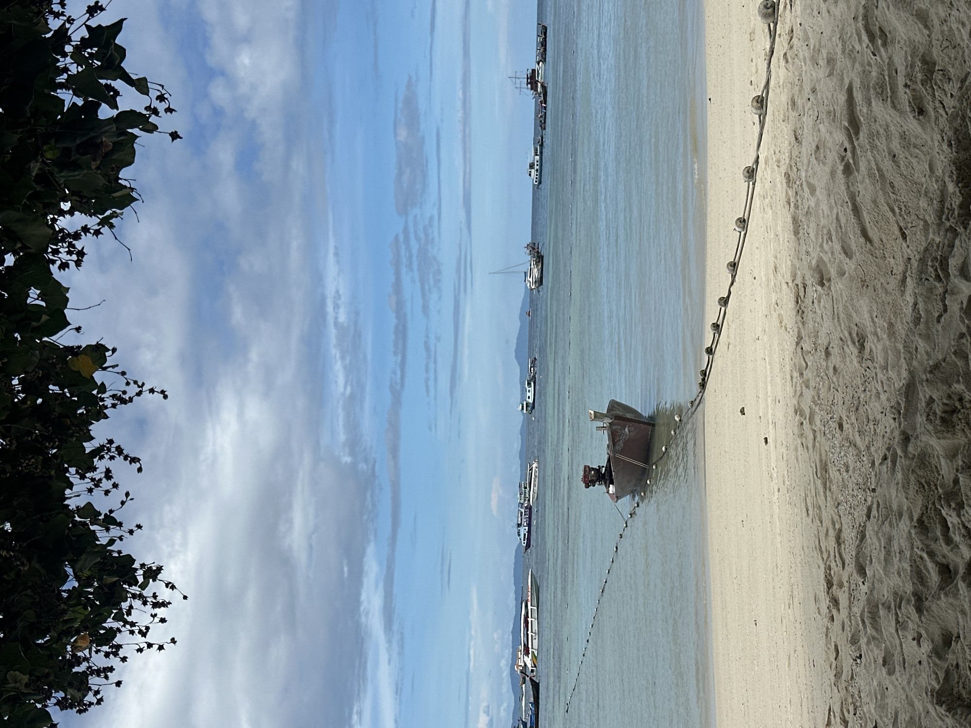 This image depicts a serene beach scene in Ao Nang, Thailand. In the foreground, the sandy beach transitions into clear, calm turquoise waters. A small, brown wooden boat is anchored close to the shore, tethered by a rope with buoys. The water extends to the horizon, dotted with numerous boats of various sizes, including speedboats, sailboats, and larger vessels, suggesting a popular tourist destination. The sky above is a mix of blue and white clouds, with the silhouette of distant islands visible in the background. Overhead, the branches of a leafy tree frame the top of the image, casting subtle shadows. The overall atmosphere is peaceful and idyllic, characteristic of a tropical coastal town.