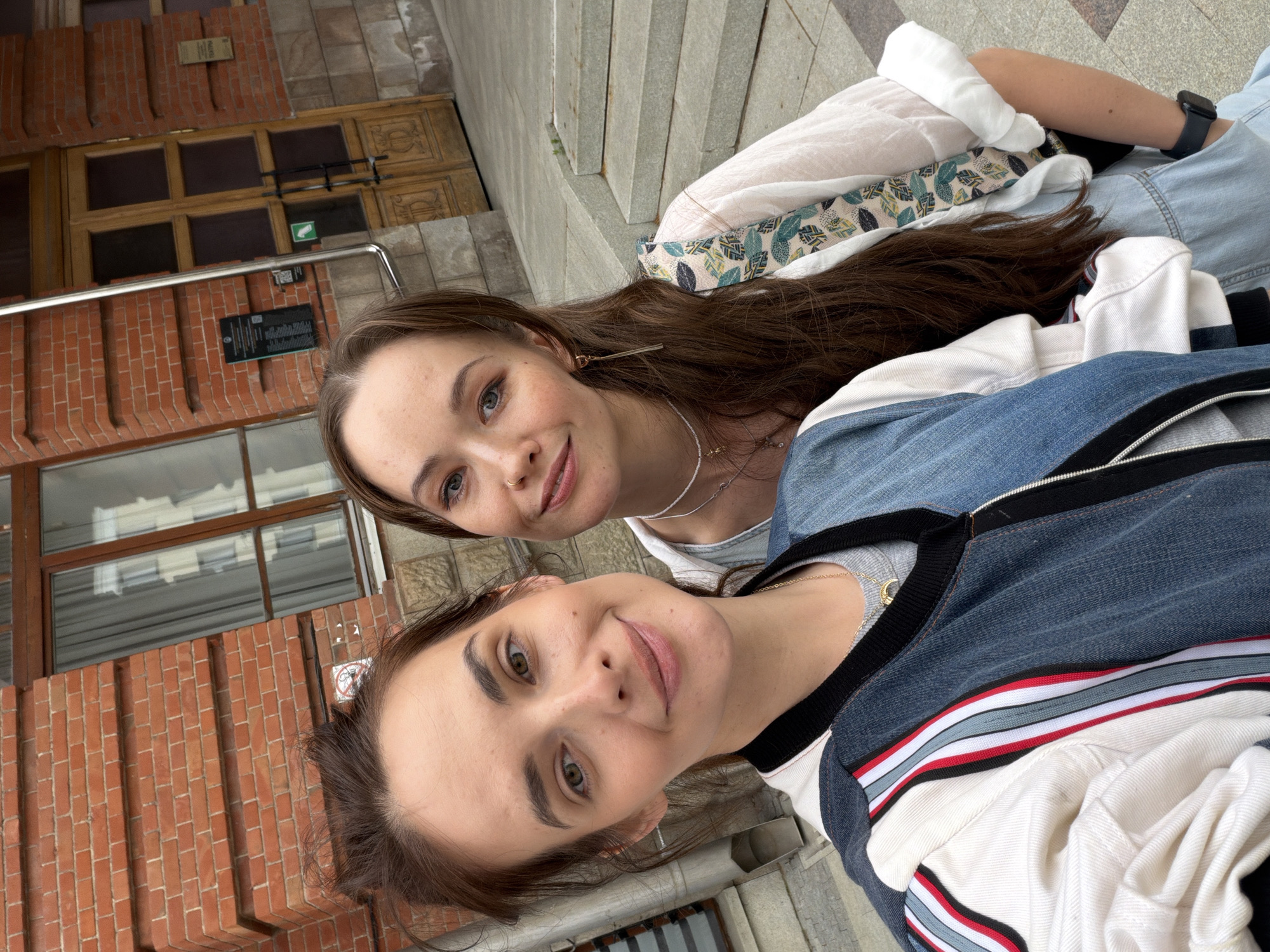 The image is a selfie of two young women outdoors. One woman is in the foreground, looking directly at the camera with a smile. She has brown hair, green eyes, and is wearing a grey t-shirt and a denim jacket with white and red stripes. The second woman is behind her, also looking at the camera with a smile. She has long brown hair, blue eyes, and is wearing a white sheer top with a decorative trim and a denim jacket.
They are standing in front of a brick building with large wooden doors and windows. There are stone steps behind them, suggesting an outdoor public space or the entrance to a building. The lighting indicates it is daytime, and the overall atmosphere seems casual and happy.
One visible sign on the building has Cyrillic text. There is also a small green sign with a white symbol on it, possibly indicating a direction or rule. The clothing of the subjects and the architecture suggest a European setting, specifically Ufa, Russia.