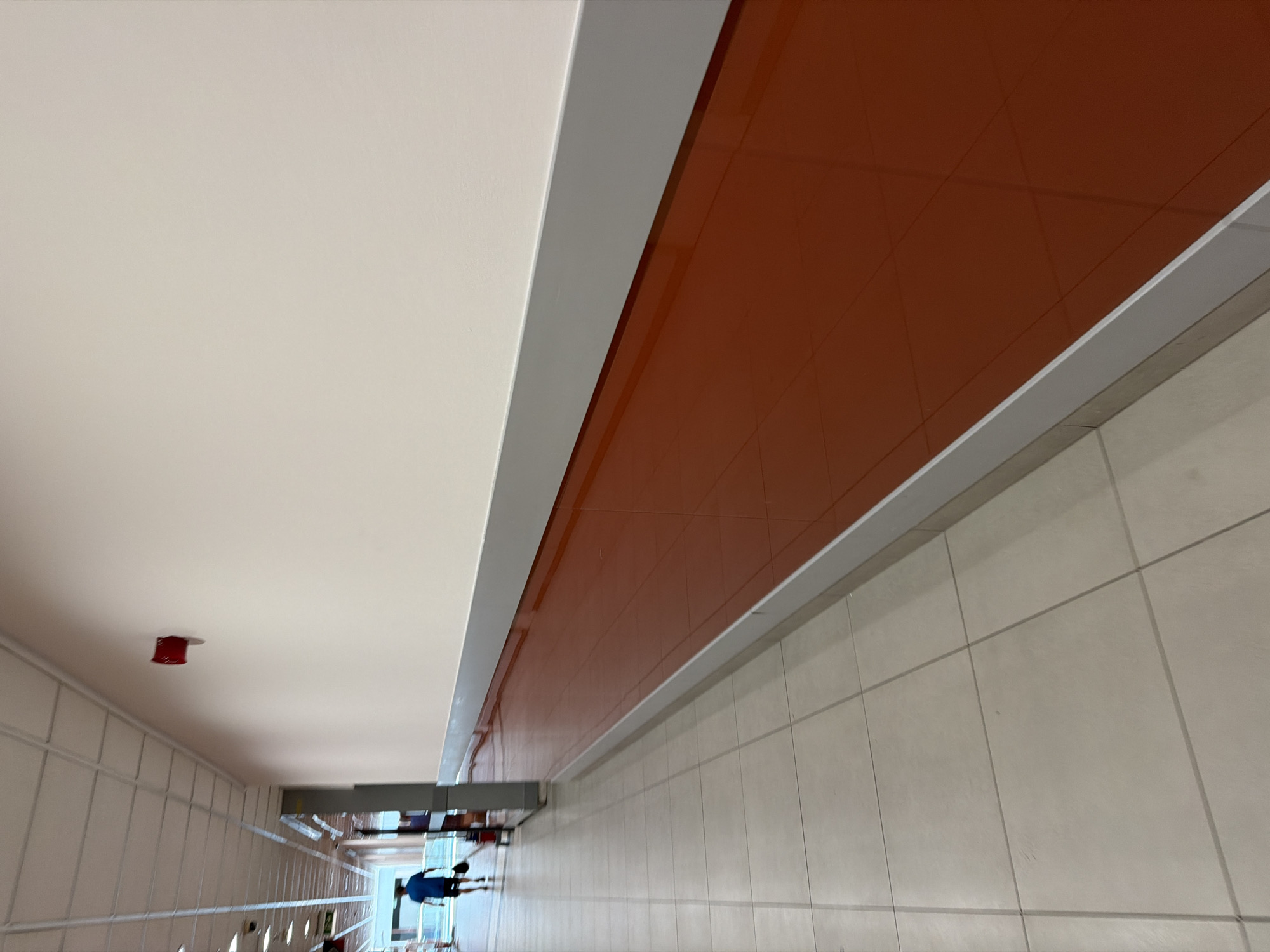 This image depicts a long, brightly lit corridor, likely in a commercial or public building. The corridor features a light-colored tiled floor and a smooth, pale-colored wall. Along the bottom of the wall runs a prominent band of polished, dark orange material, capped by a silver trim. A white ceiling with fluorescent lighting is visible at the top. In the mid-ground, a person wearing a dark blue t-shirt and shorts walks away from the camera, carrying a black bag. They are in front of a tall, grey fixture, possibly a display or shelving unit. Further down the corridor, another red object is mounted high on the wall, which could be a fire alarm or similar safety equipment. The overall impression is one of a clean, modern, and functional interior space. The image is not specific enough to determine its precise location in Sofia, Bulgaria, but it is consistent with the interior of a shopping mall, airport, or large retail establishment.